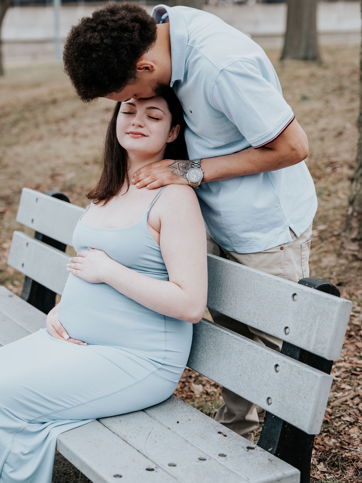 Pregnant woman in light blue dress sits on park bench as partner kisses her head during maternity photoshoot.