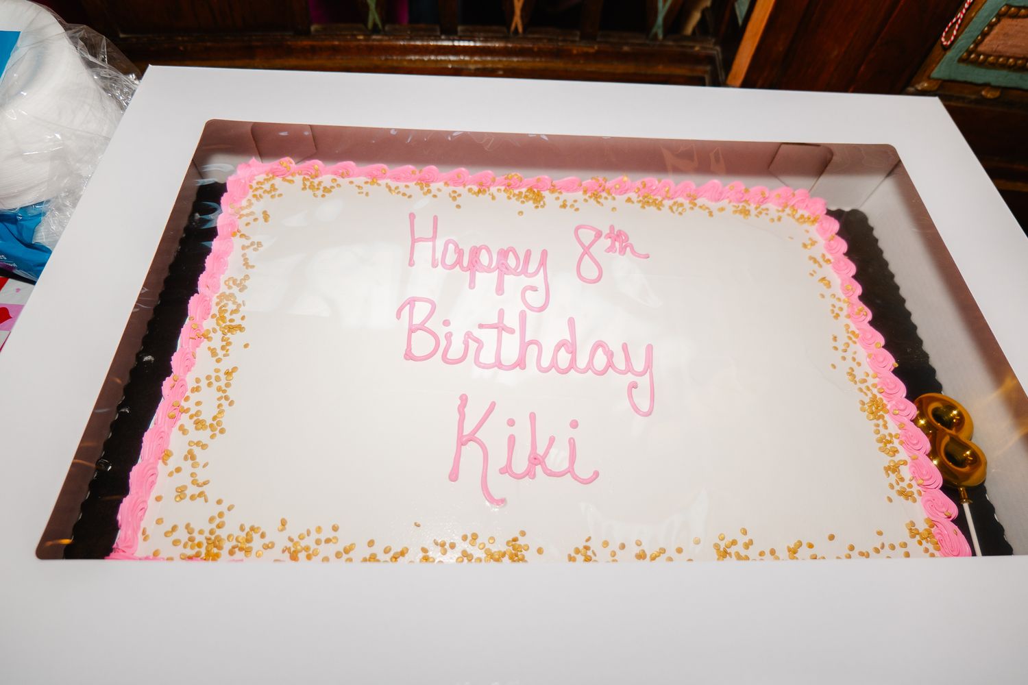 White sheet cake with pink frosting reading Happy 8th Birthday Kiki with gold star sprinkles.