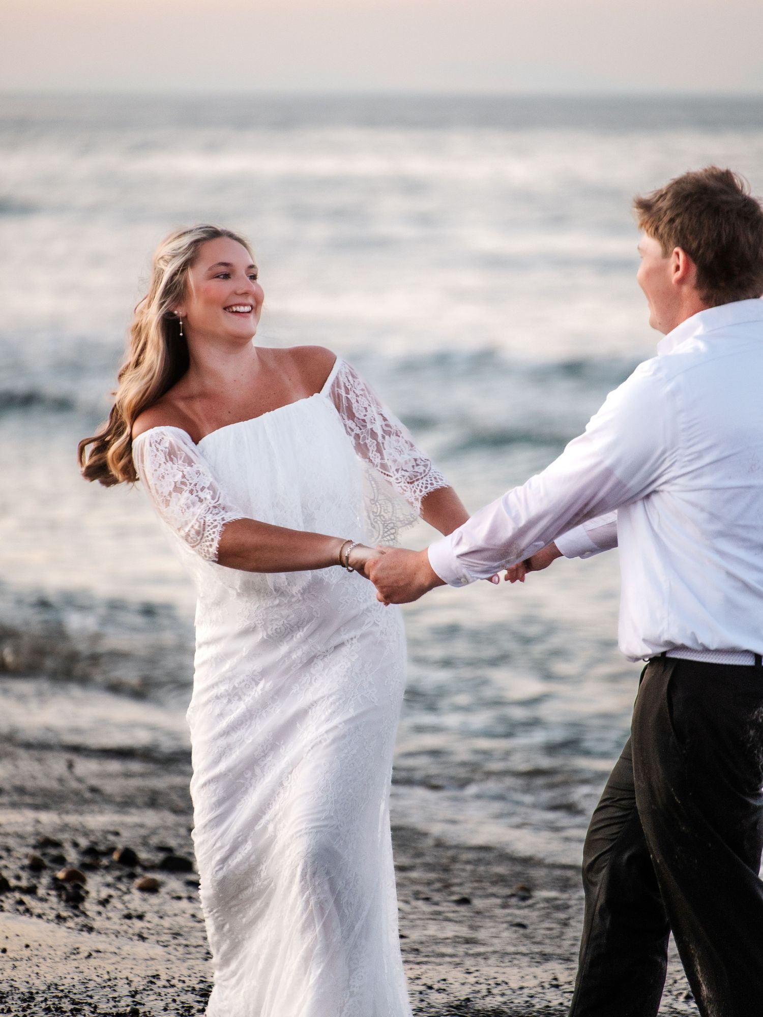 Smiling bride in white lace off-shoulder gown dancing with groom on beach at sunset.