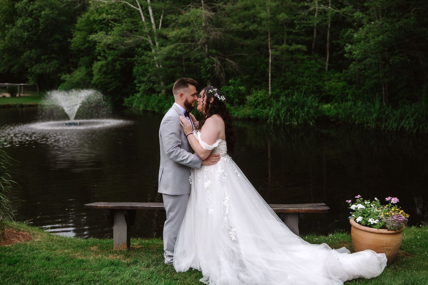 Bride and groom share a romantic kiss near a serene pond with a fountain, surrounded by lush greenery on their wedding day.