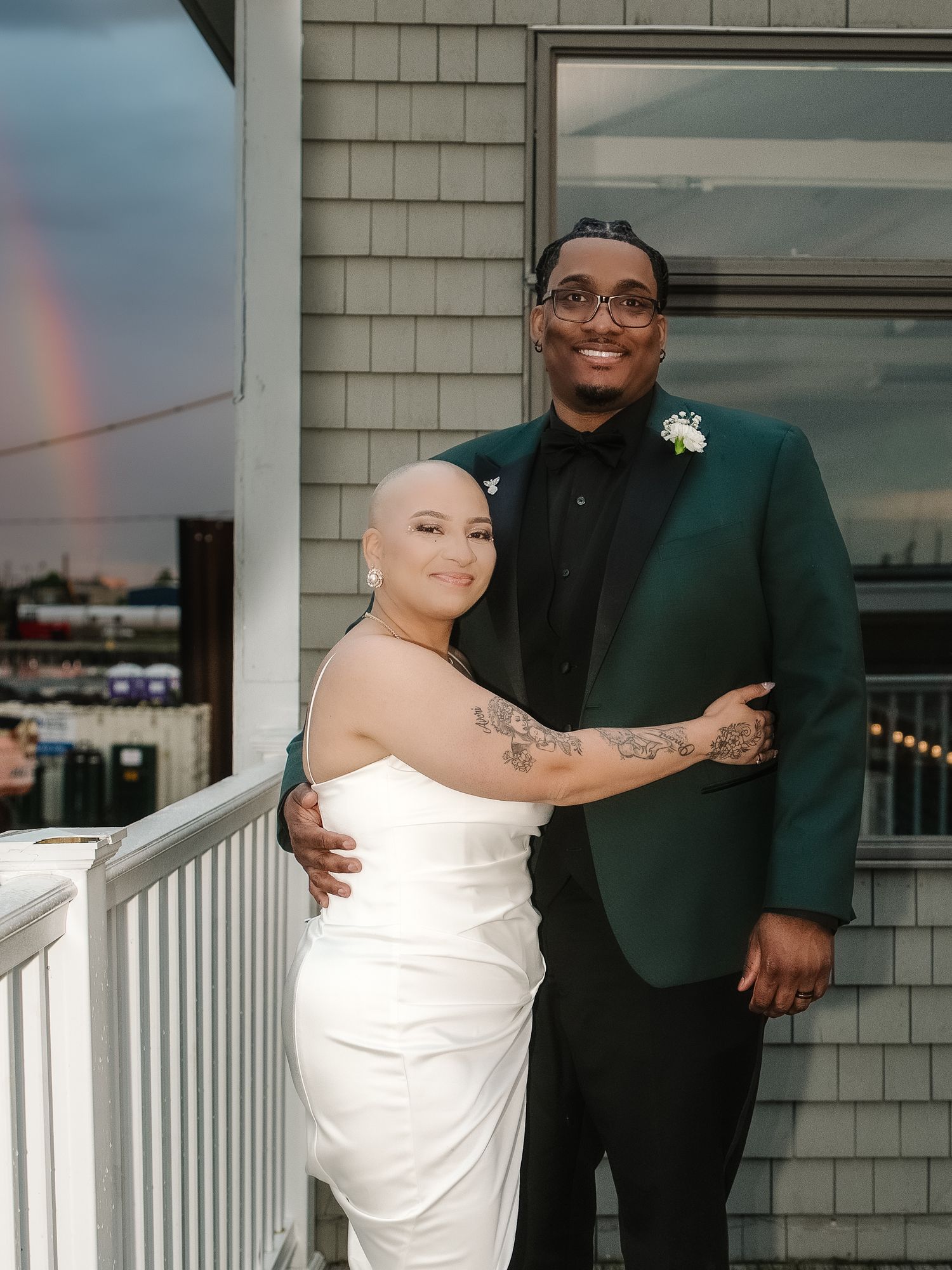 Couple in wedding attire poses on balcony, bride in white gown, groom in green suit, rainbow visible in background.