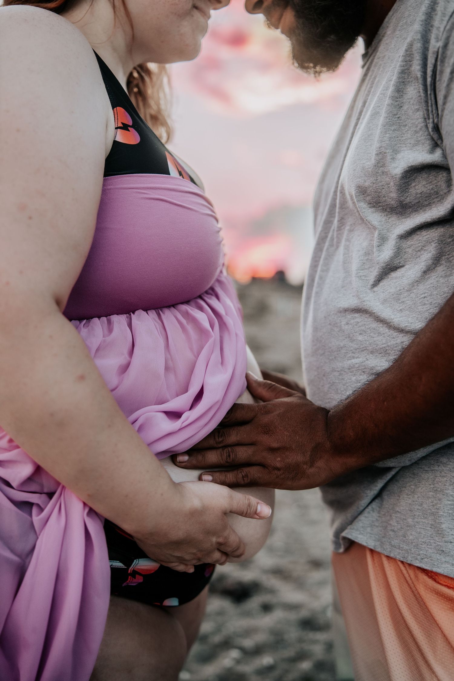Couple tenderly cradles pregnant belly on beach, woman in purple maternity gown, partner in gray shirt.