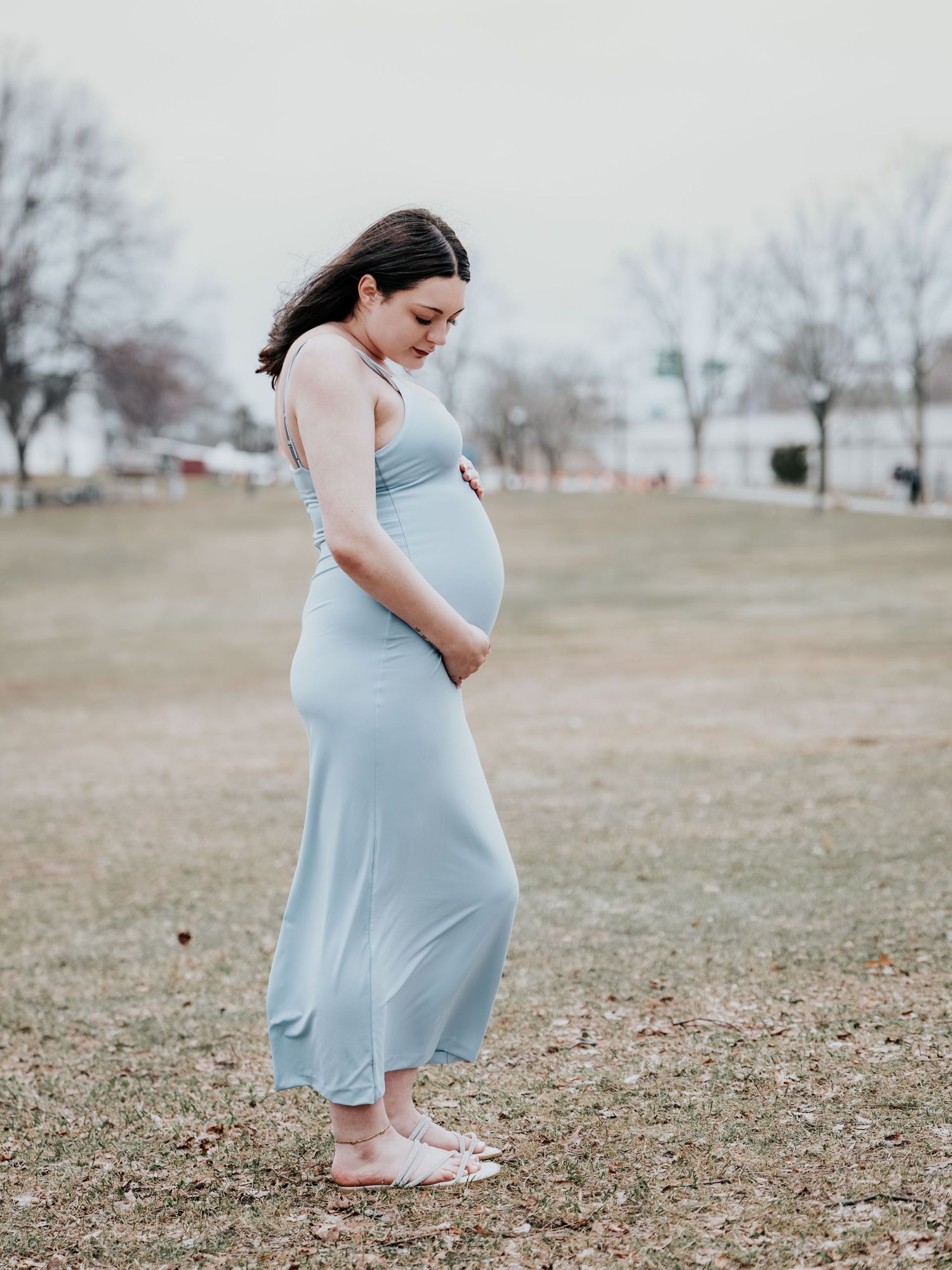 Pregnant woman in light blue maxi dress cradling baby bump outdoors in a park during maternity photo session.