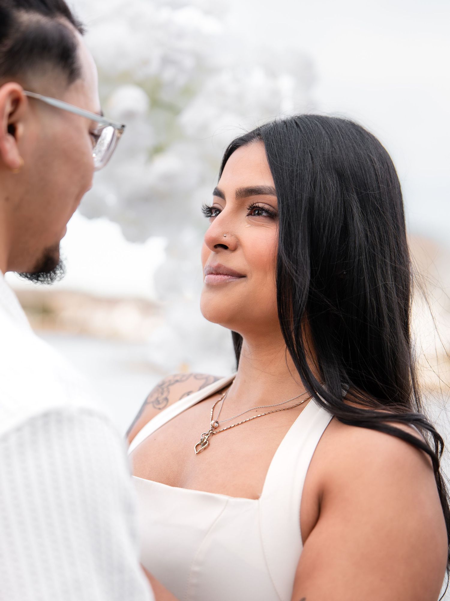 Woman in white dress gazing lovingly at her partner during an outdoor proposal with white flowers.