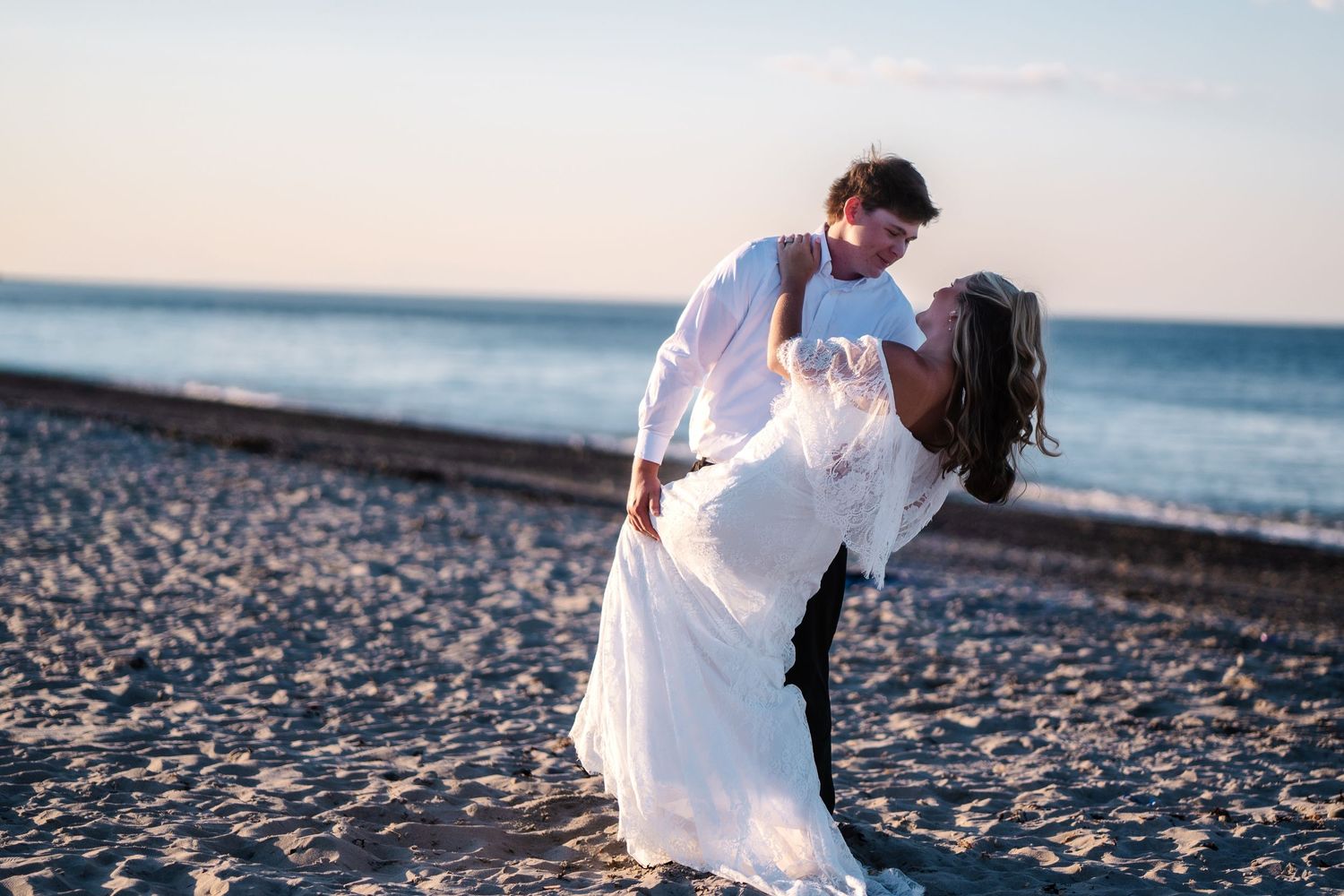 Groom lifting bride in white lace dress on sandy beach at sunset with ocean waves in background.