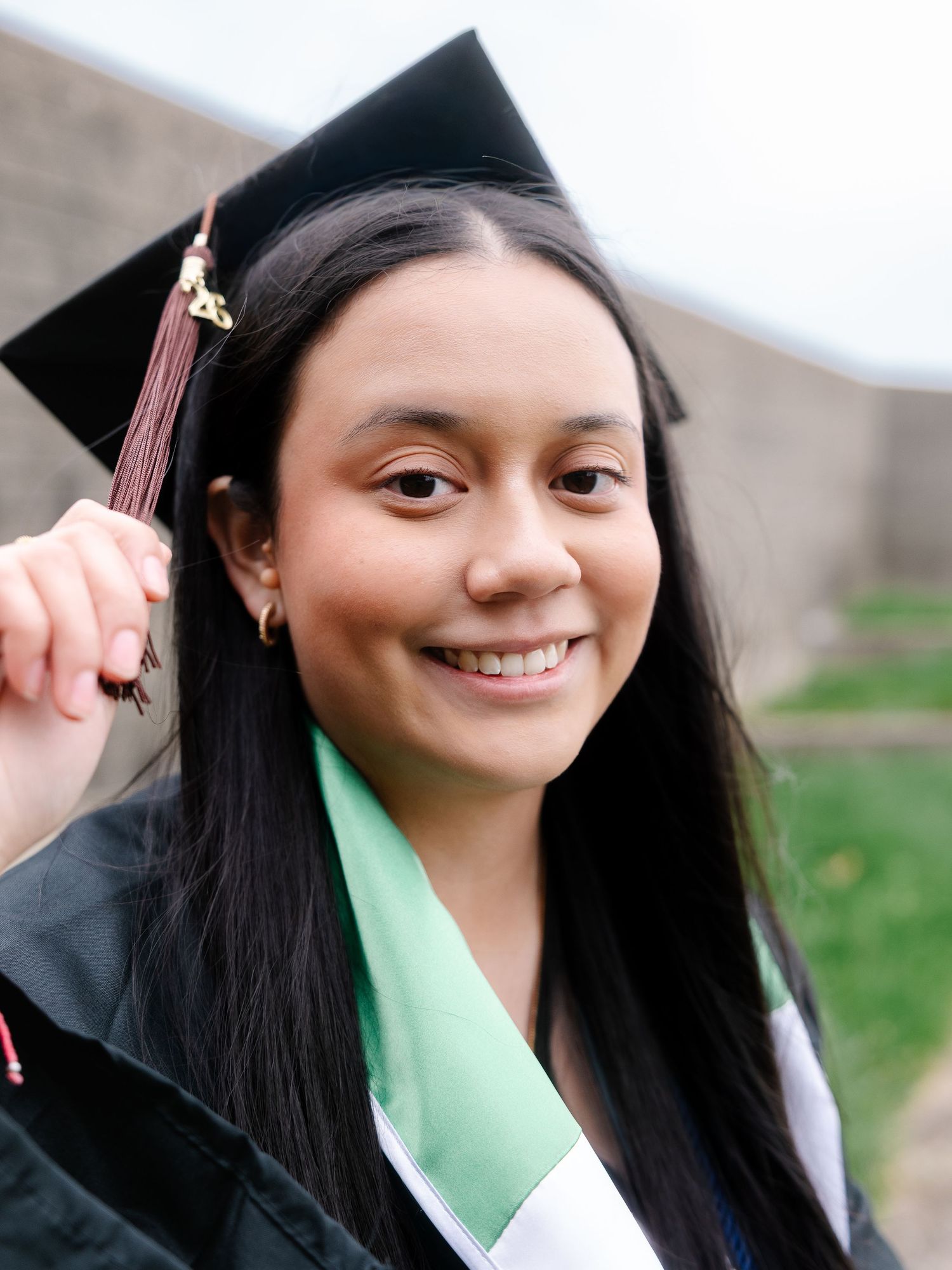 Close-up of smiling graduate holding graduation cap tassel wearing green stole.