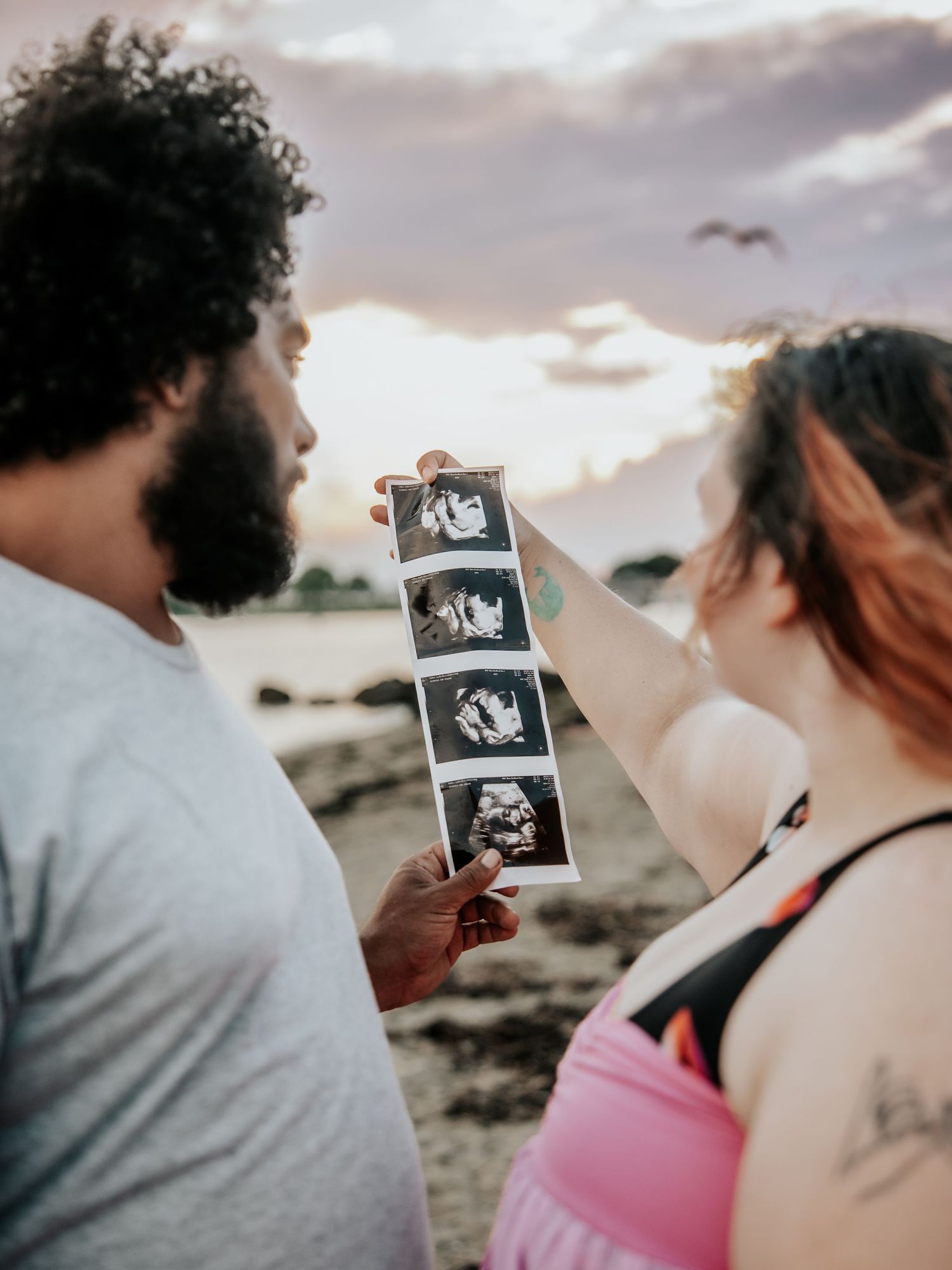 A couple looks at ultrasound photos together on a beach at sunset, celebrating their pregnancy announcement outdoors.