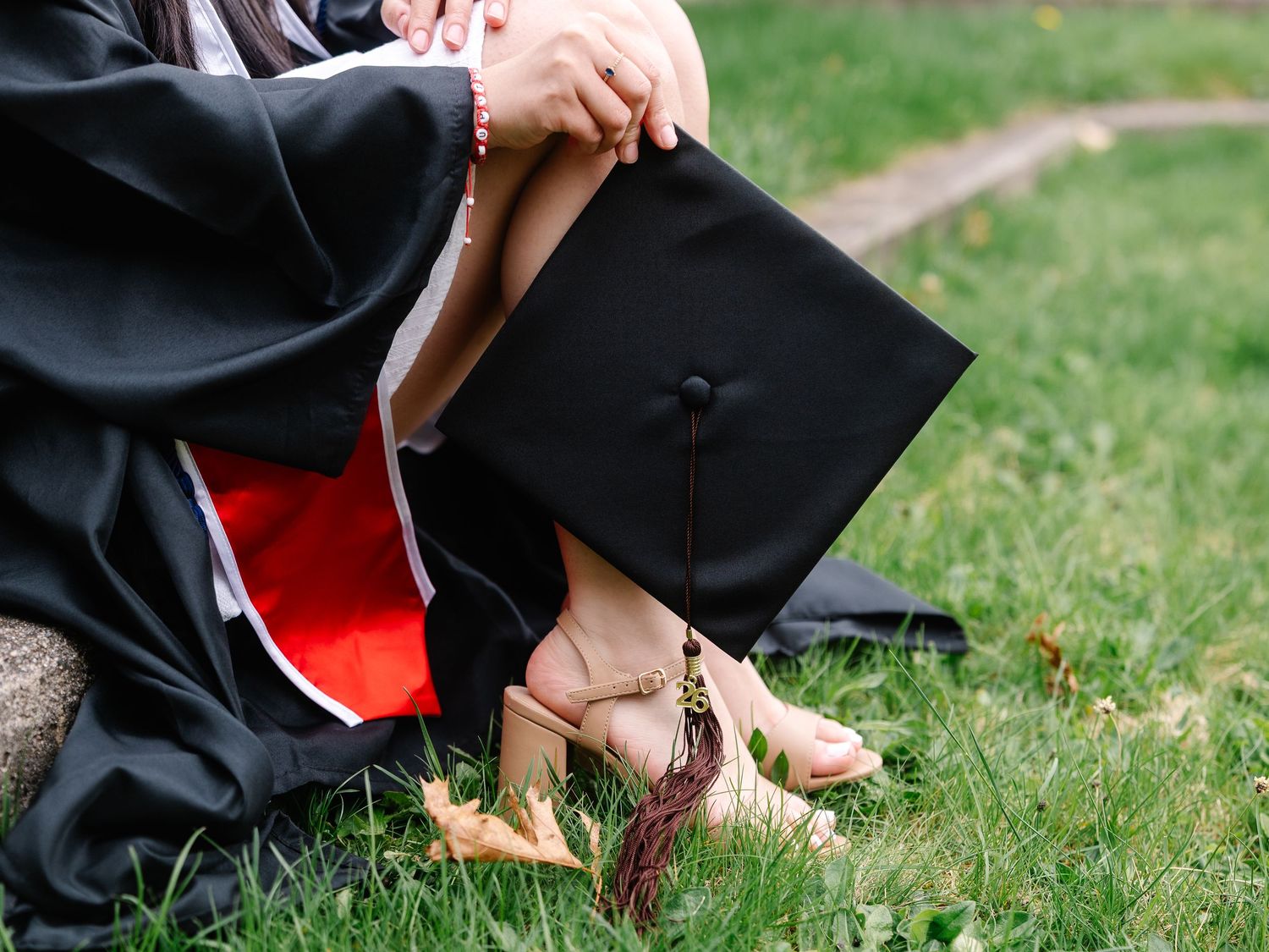 Close-up of graduate in black robe sitting on grass holding graduation cap.
