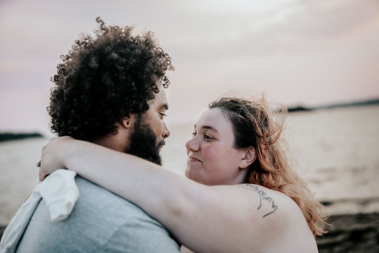 Romantic couple embracing near water at sunset, woman with tattoo hugging curly-haired man on beach.