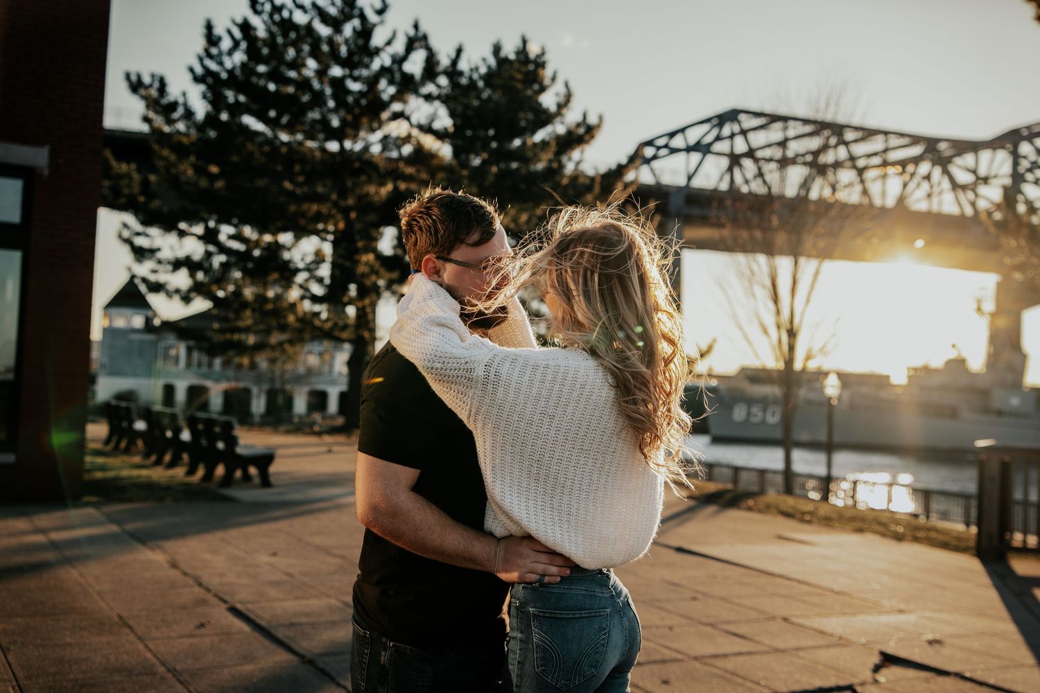 Couple embracing and kissing at sunset near a waterfront with a steel bridge in the background.