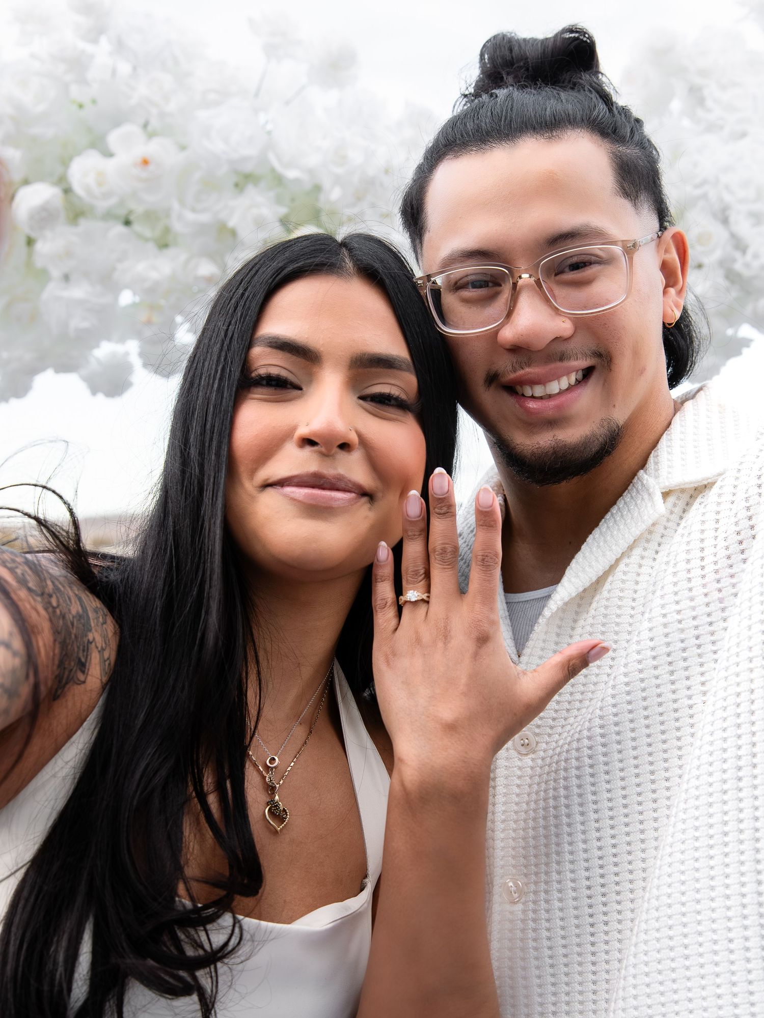 Couple smiles at camera showing off engagement ring with white floral backdrop.