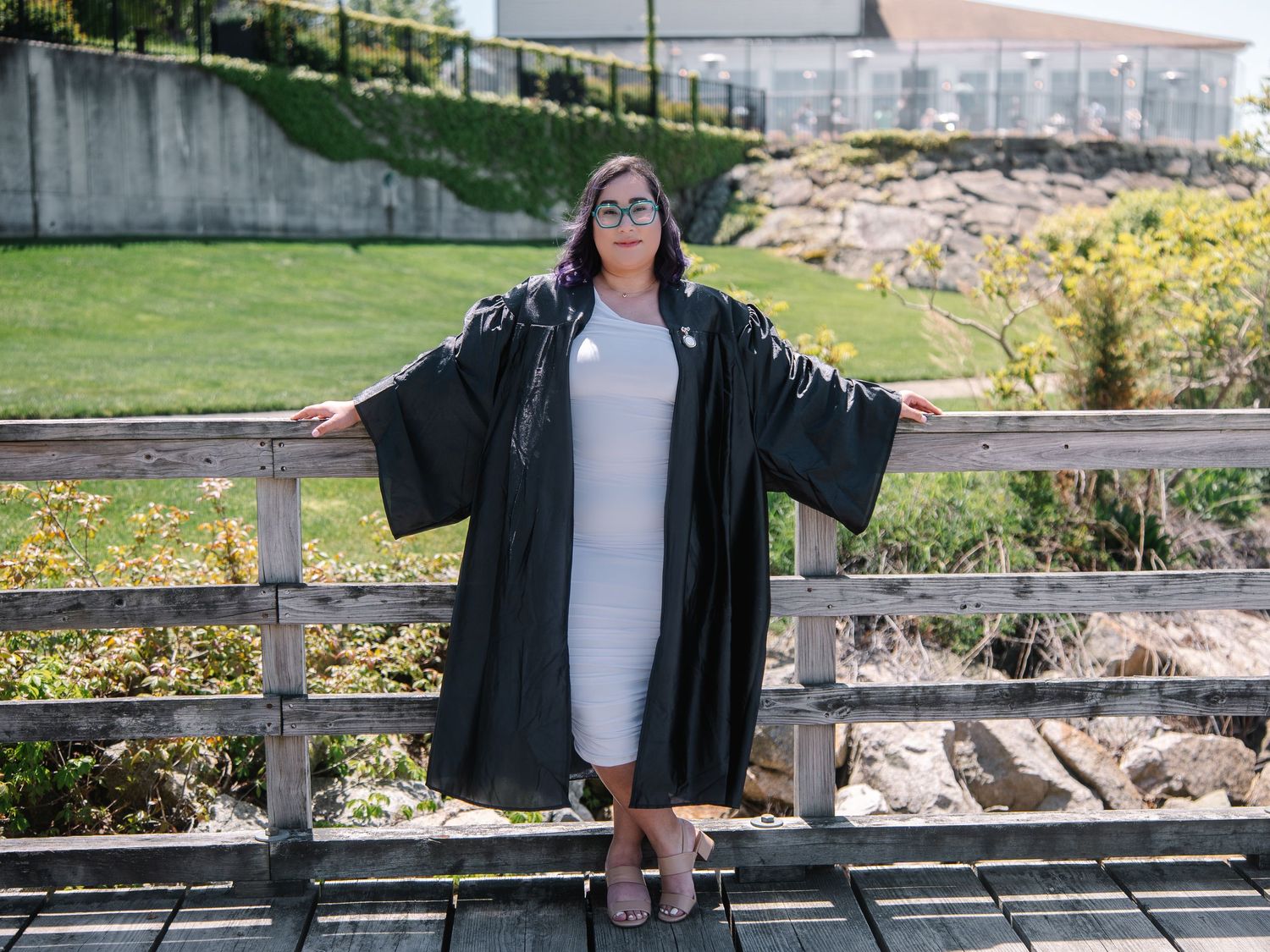 Graduate in black robe posing on wooden bridge wearing white dress and glasses outdoors.
