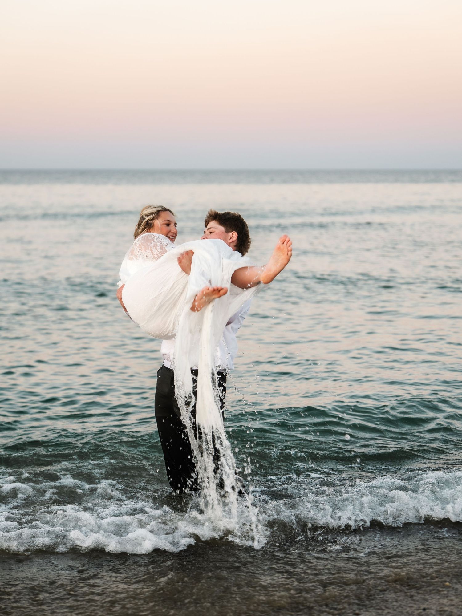 Groom lifting bride in white lace dress while standing in ocean waves at sunset.