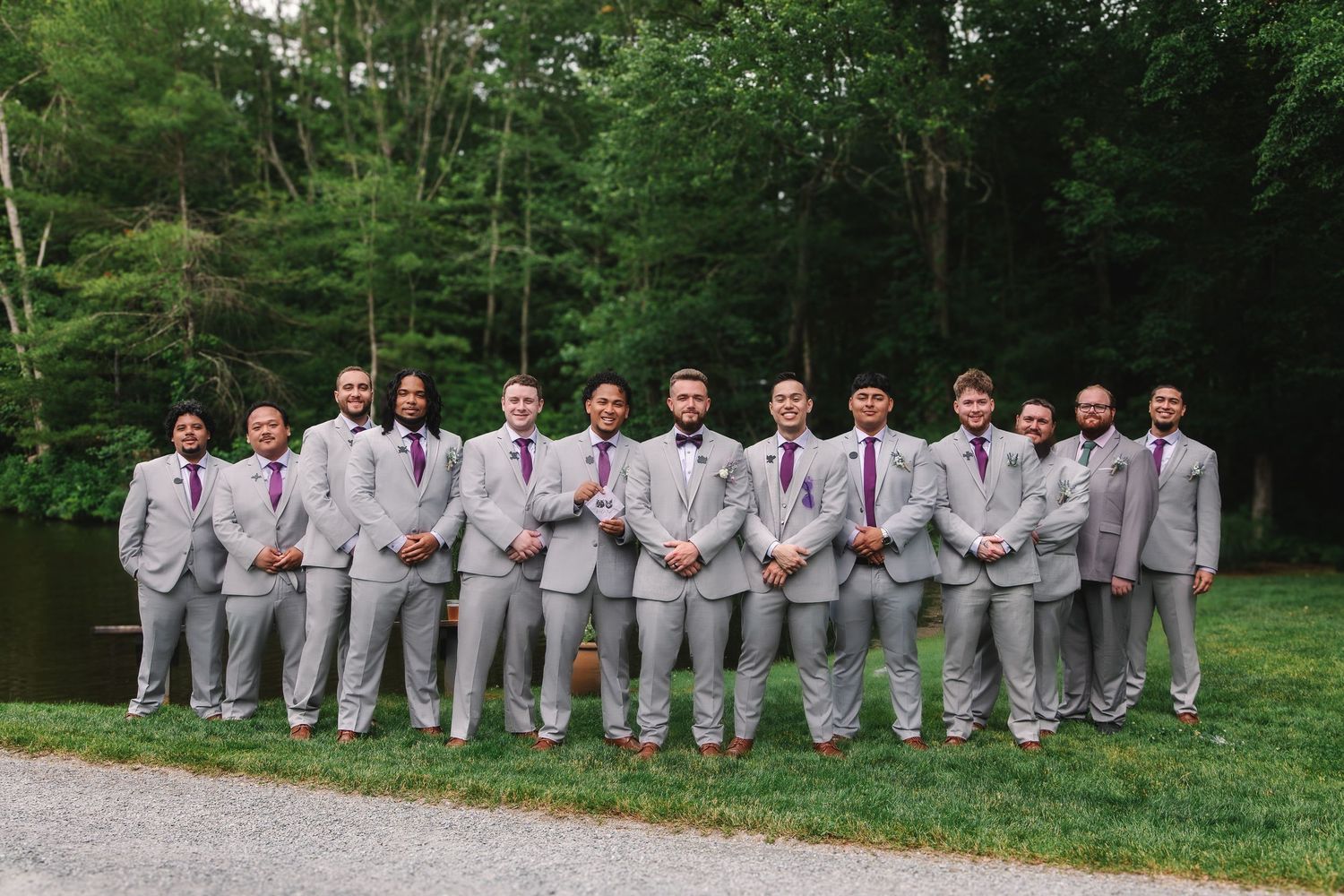 Groomsmen in gray suits with purple ties posing outdoors near a pond with lush green trees in the background.