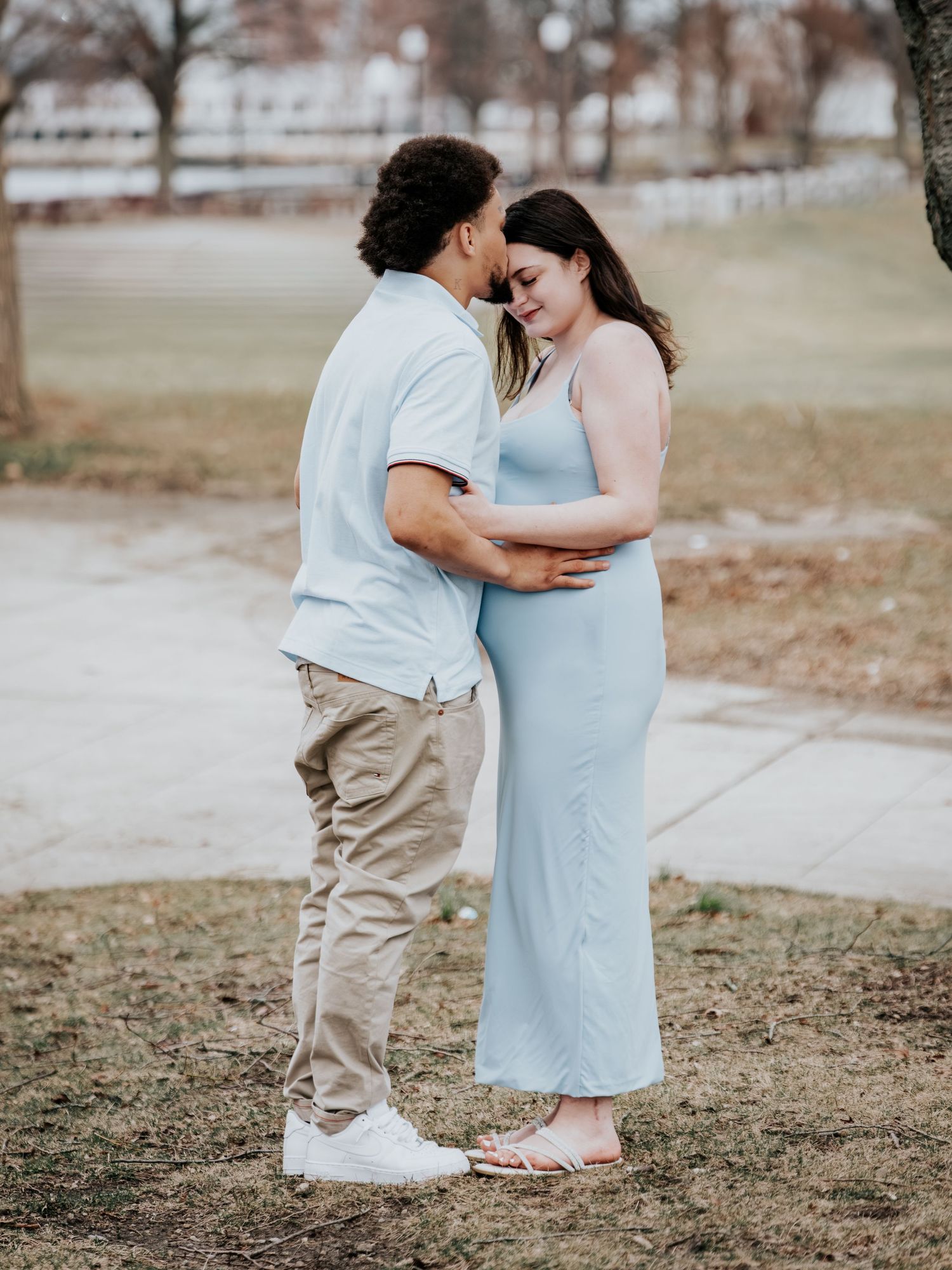 Expecting couple embracing tenderly outdoors, man in light blue polo and khakis holding pregnant partner in blue dress.