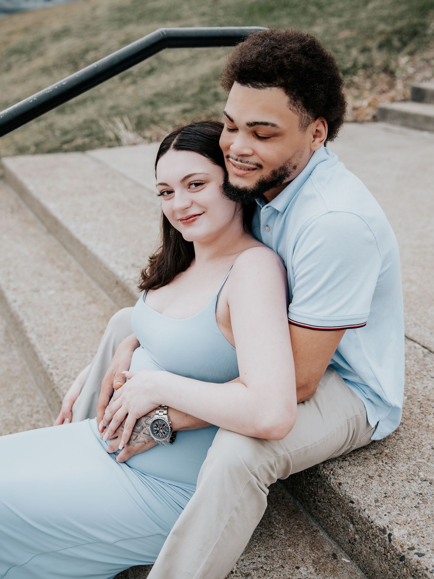 A couple sits on stone steps, the woman in a blue maternity dress leaning against her smiling partner in a light blue polo.