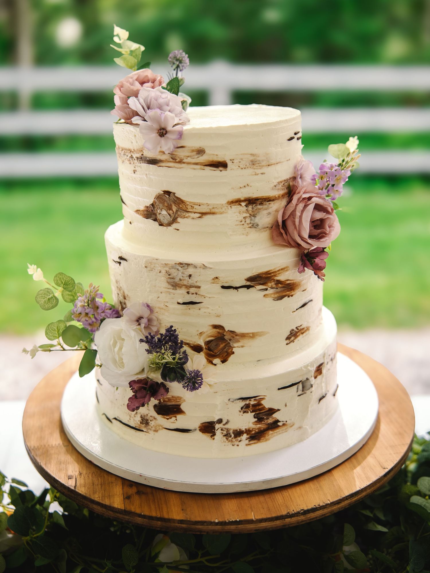 Three-tier rustic wedding cake with birch bark texture, adorned with pink, purple, and white flowers on a wooden stand.