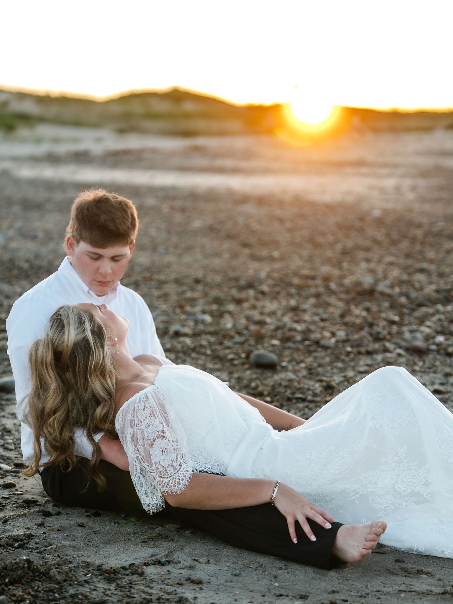 Groom in white shirt holding bride in lace wedding dress on sandy beach during golden hour sunset.