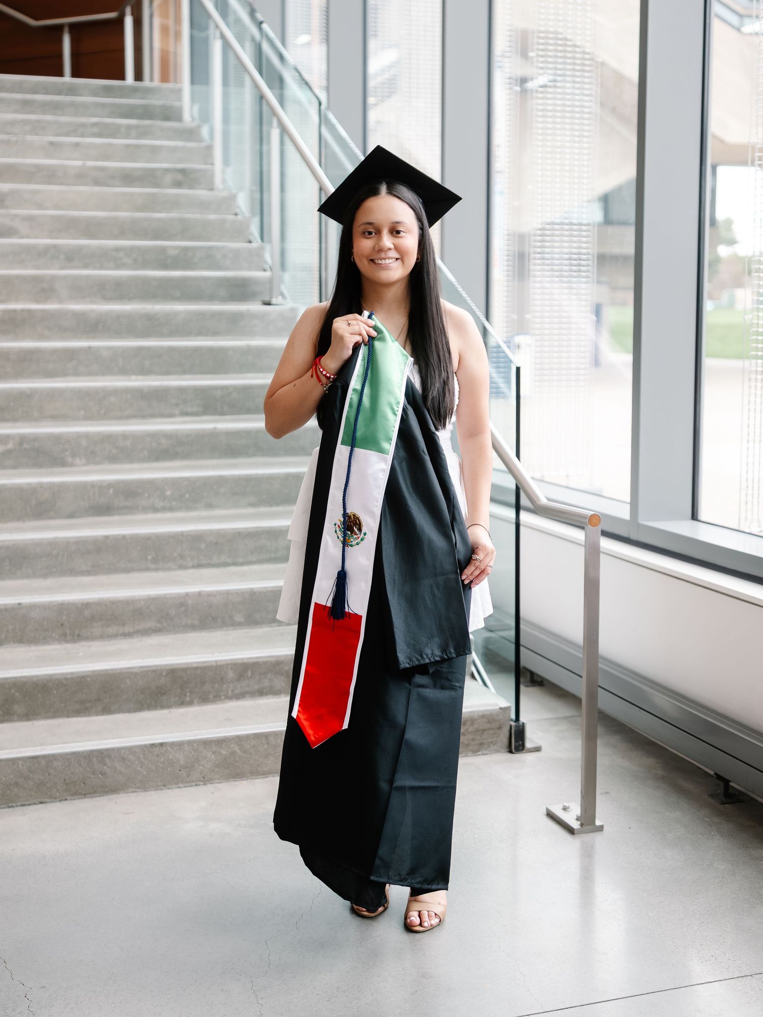 Graduate in black cap and gown proudly displays Mexican flag stole near modern staircase.