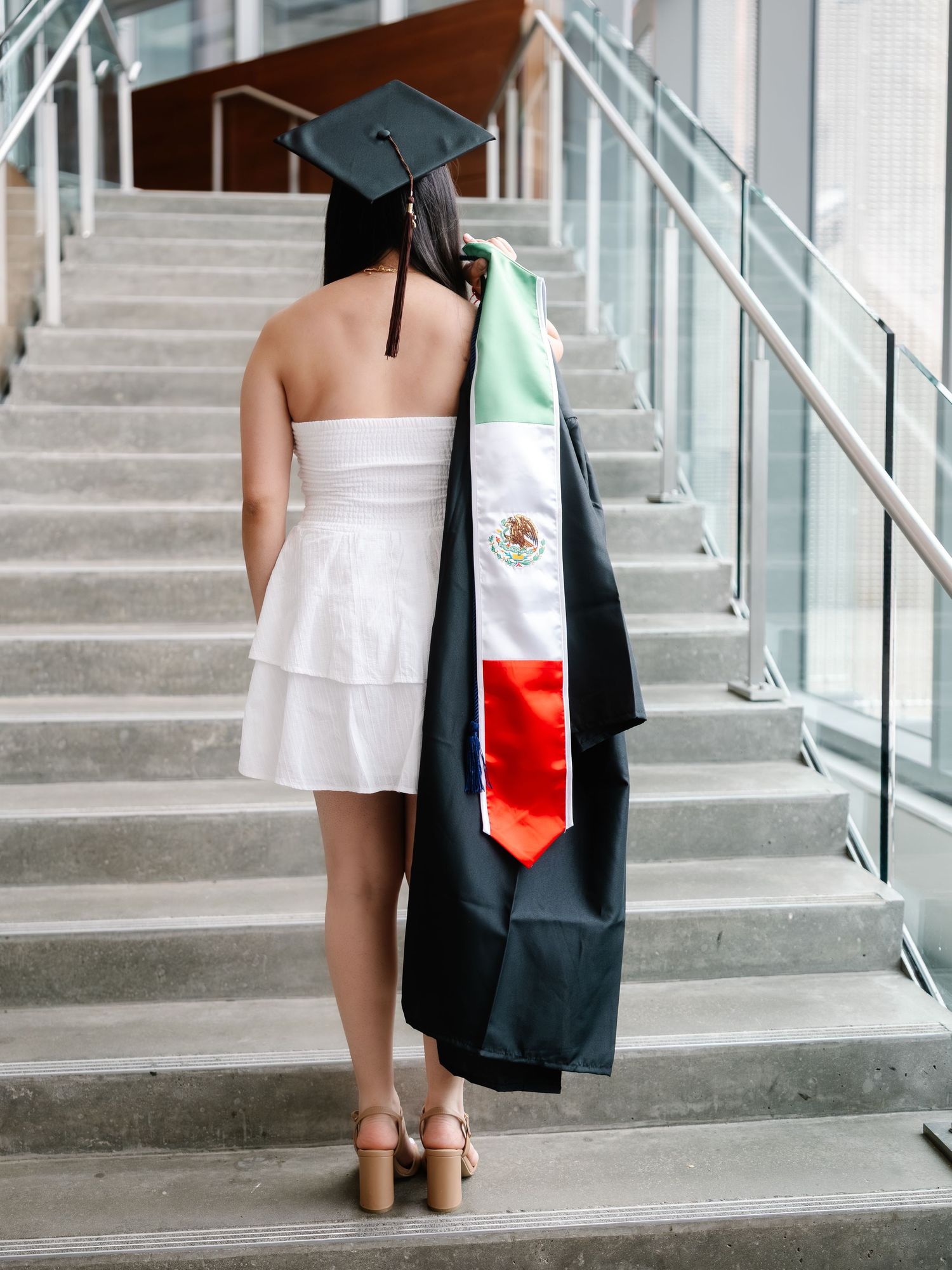 Graduate in white dress and black cap walks up stairs draped in Mexican flag stole.