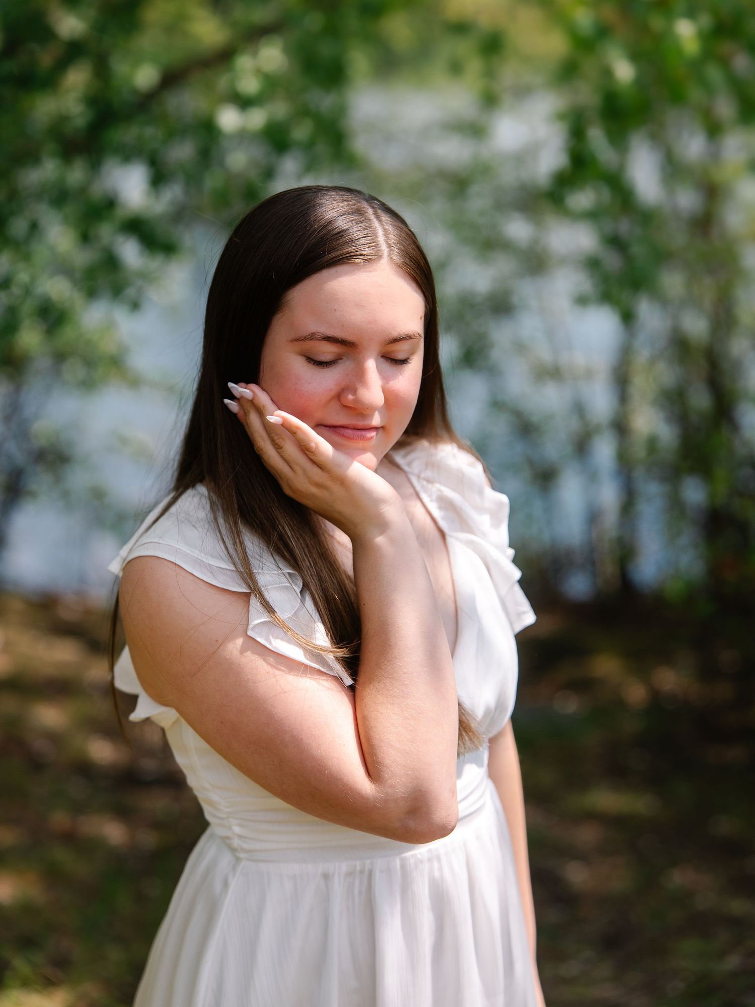 Young woman in white ruffled dress with eyes closed resting cheek on hand in outdoor setting.