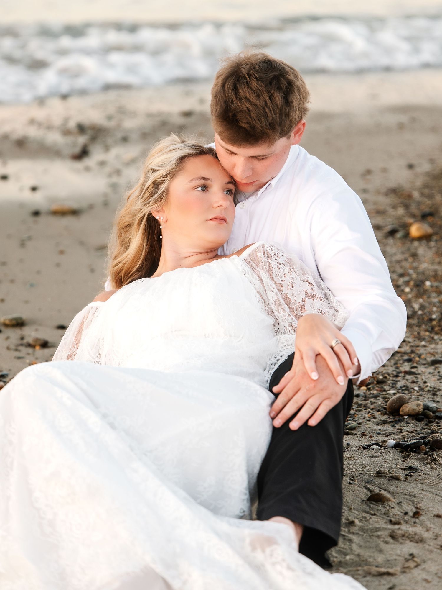 Bride in white lace gown and groom embracing tenderly on beach with ocean waves in background.