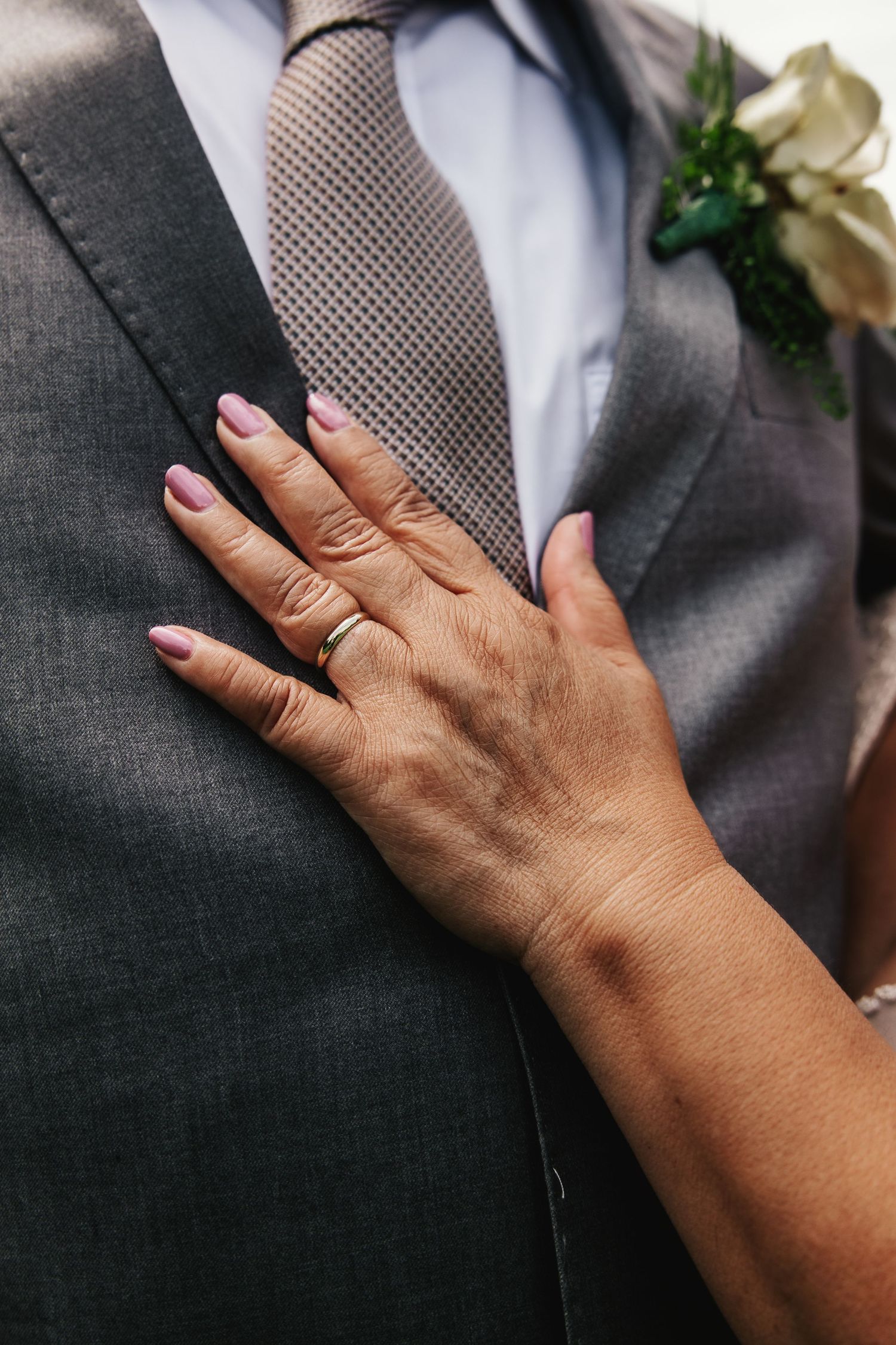 Bride's hand with wedding ring and pink nails resting on groom's gray suit jacket with boutonniere.