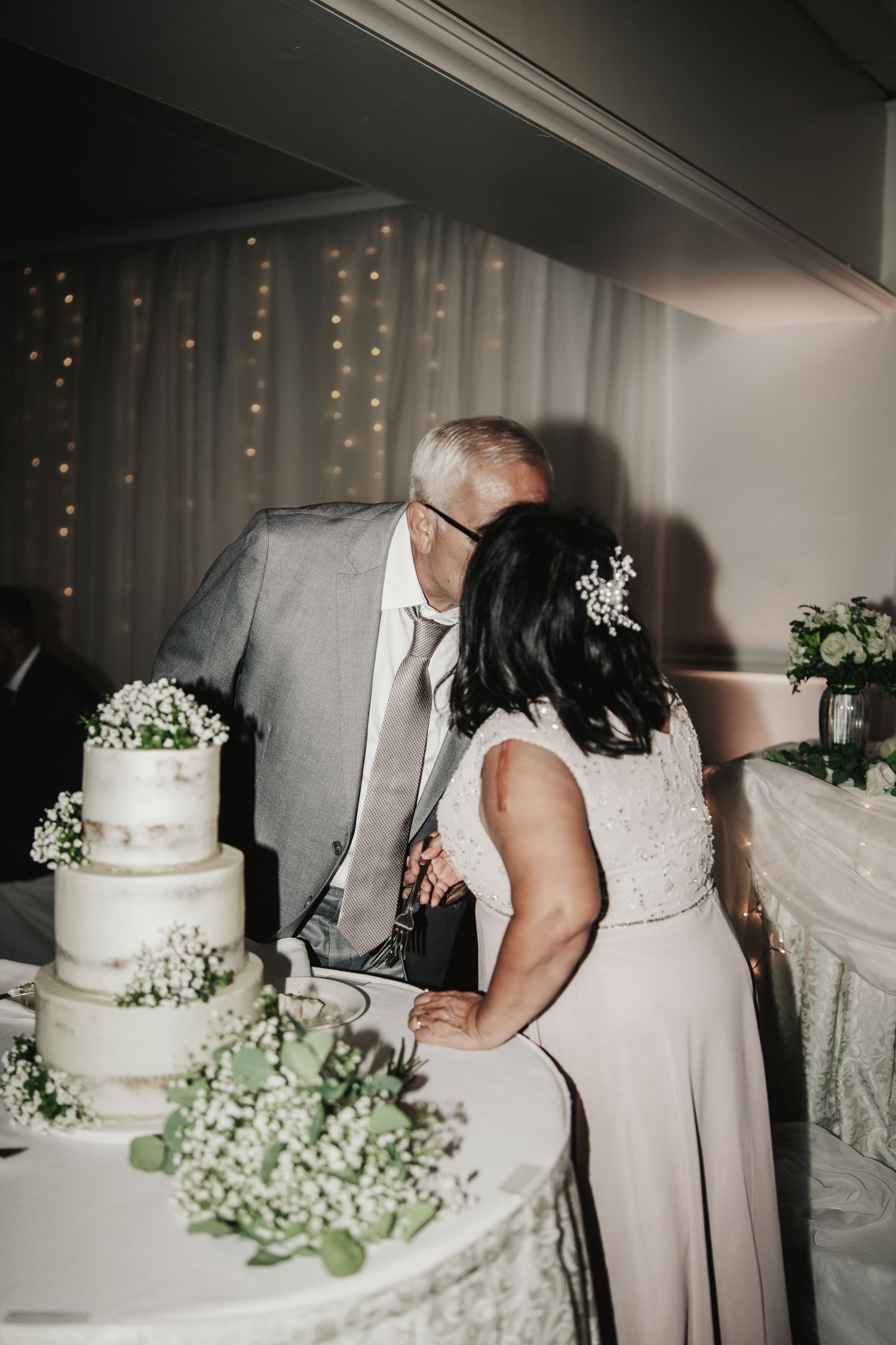 Bride and groom kissing behind a three-tier white wedding cake decorated with greenery and baby's breath flowers.