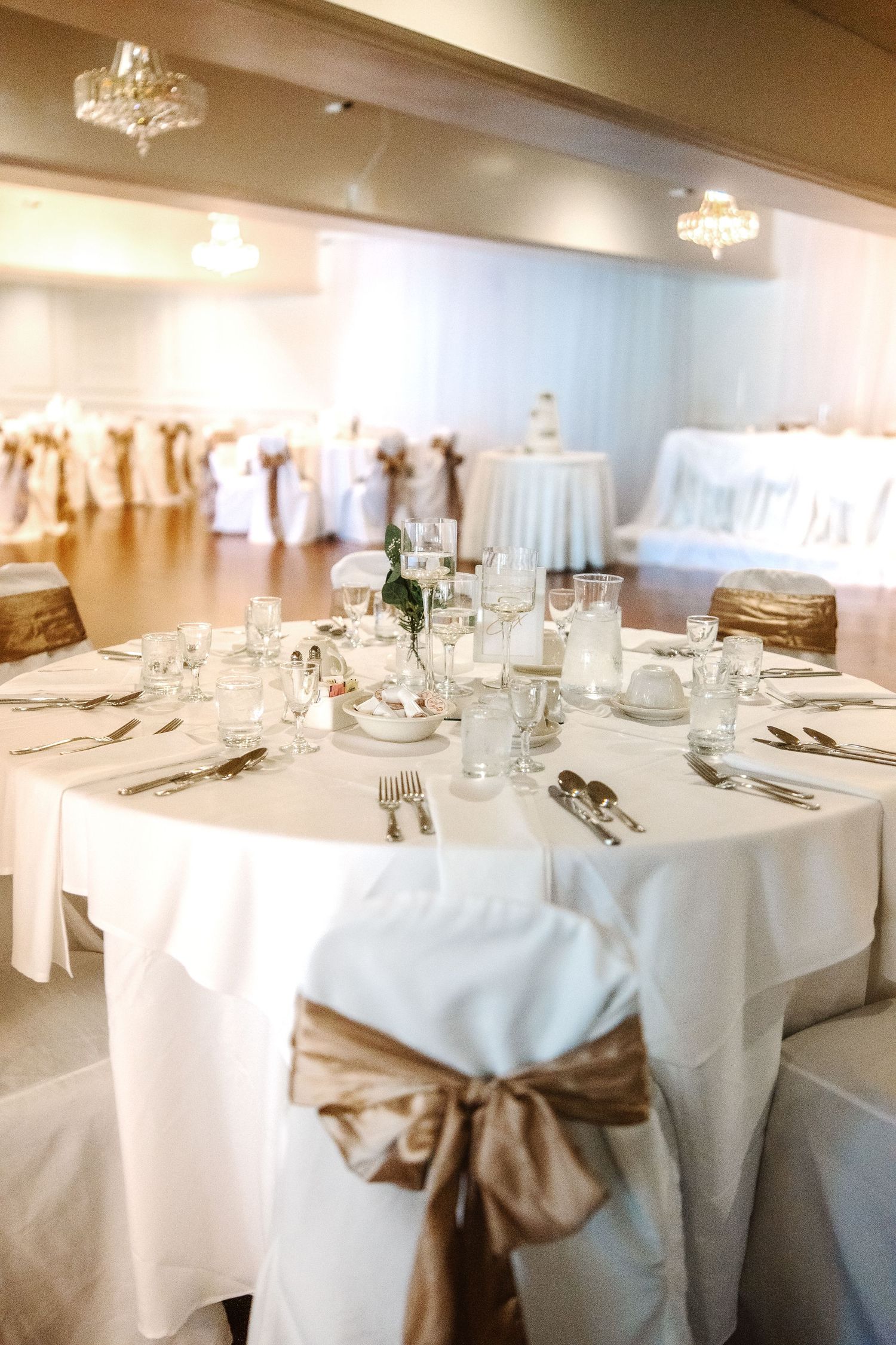 Elegant wedding reception hall with white tablecloths, gold satin chair sashes, crystal chandeliers, and formal place settings.