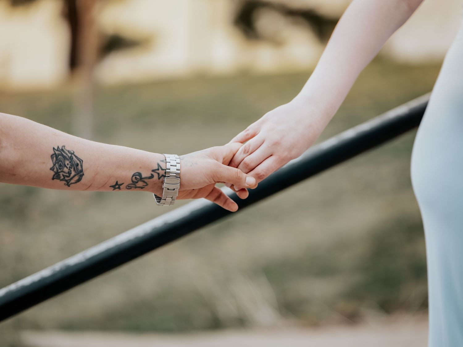 Two people holding hands over a railing, one with tattoos and a silver watch, in a romantic outdoor setting.