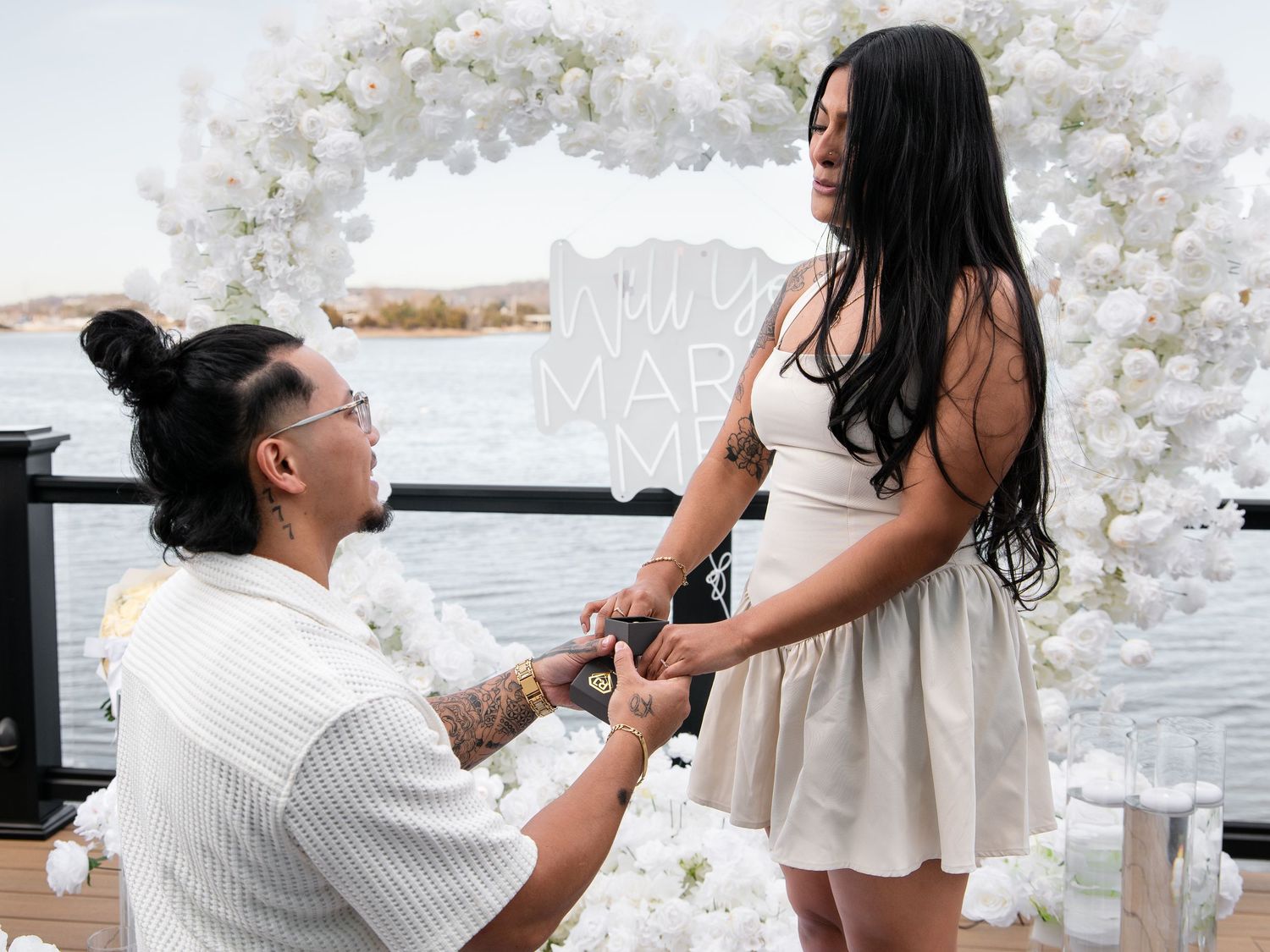 Man proposing to woman in front of floral arch near waterfront.