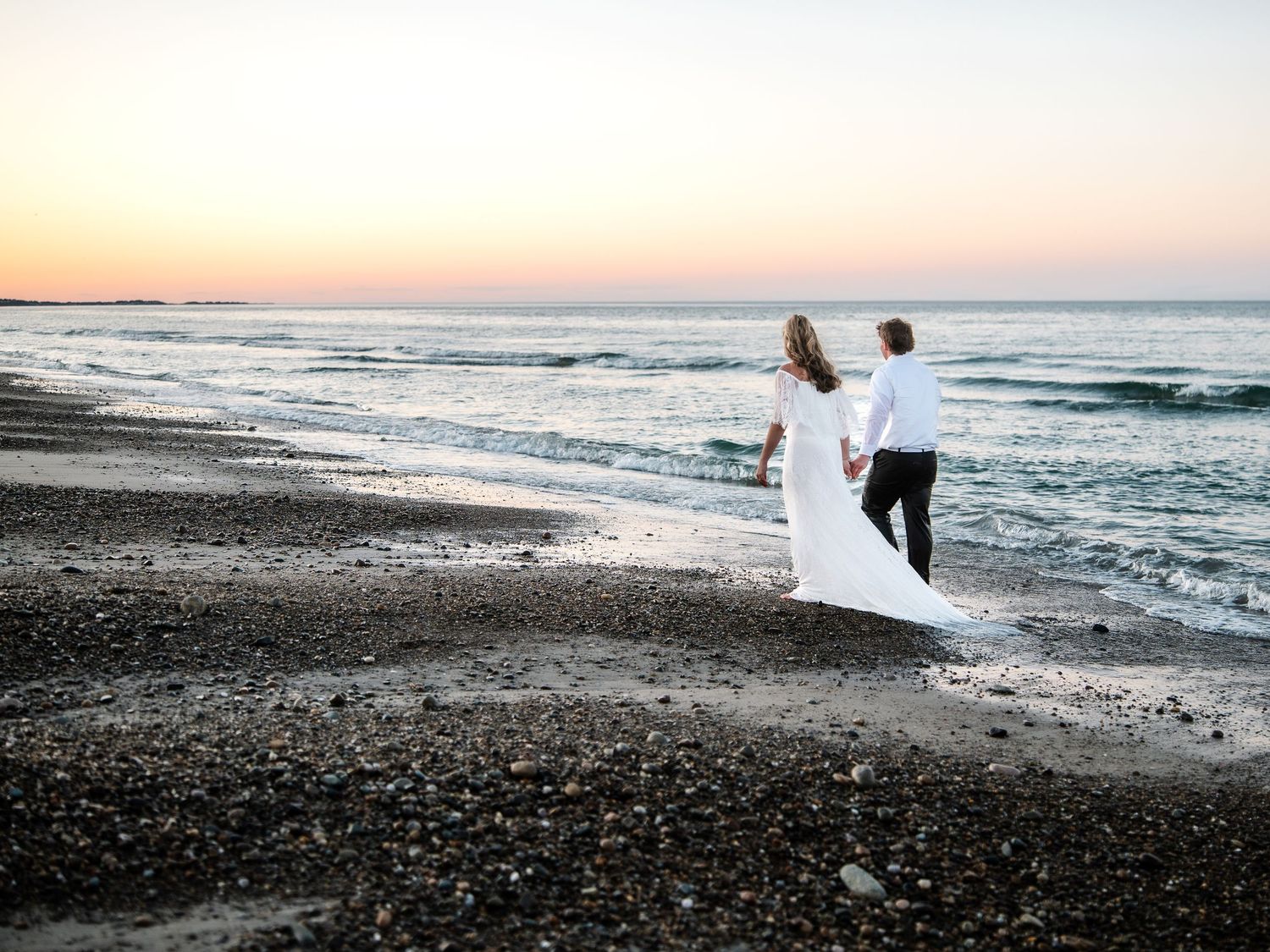 Newlyweds holding hands walking along rocky beach shoreline at colorful sunset.