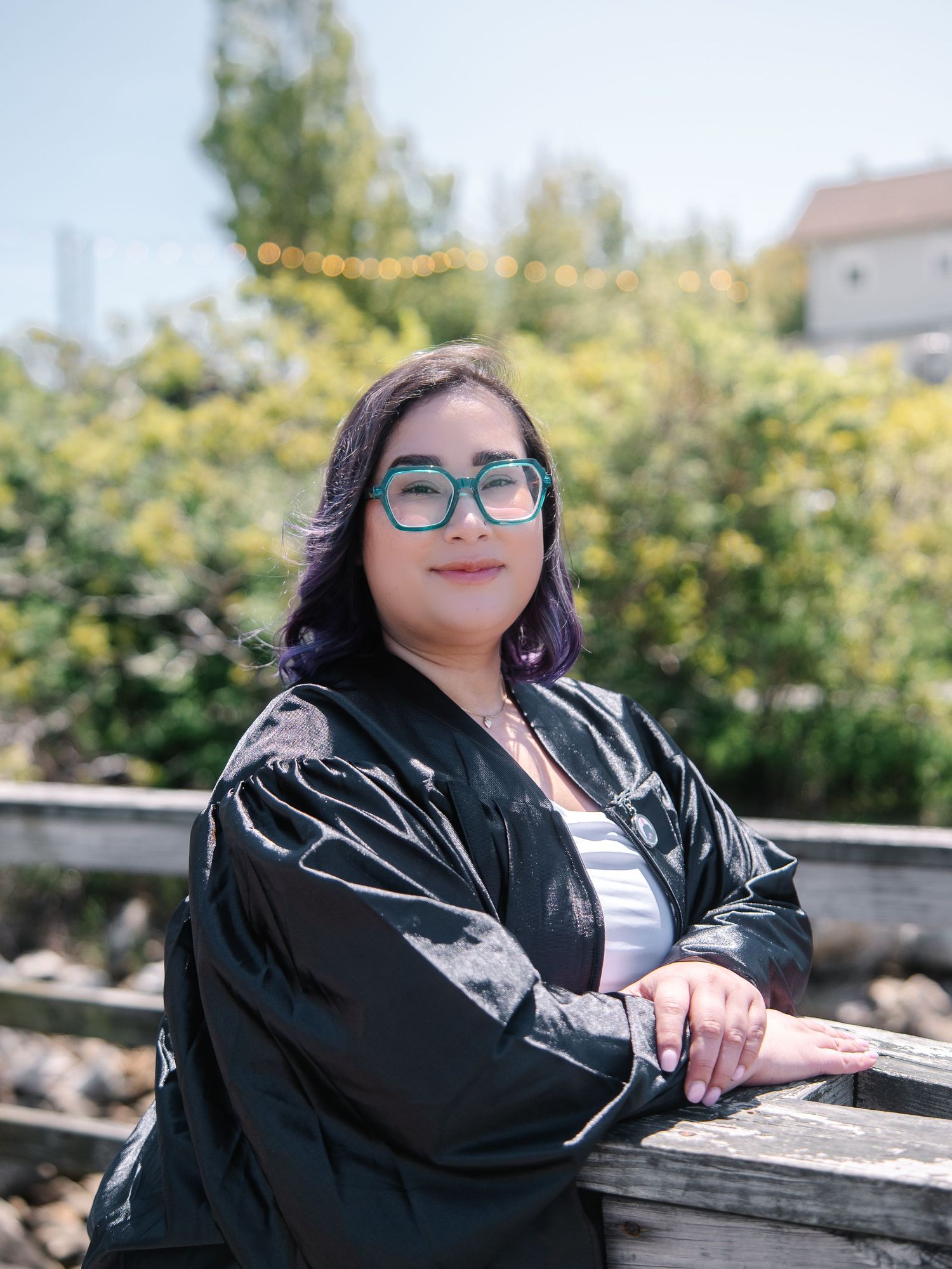 Woman in black graduation gown and teal glasses leaning on wooden railing outdoors on sunny day.