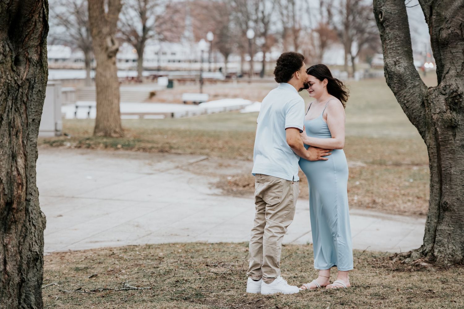 Couple sharing a tender moment in a park, woman in blue dress with visible baby bump, man in white shirt.