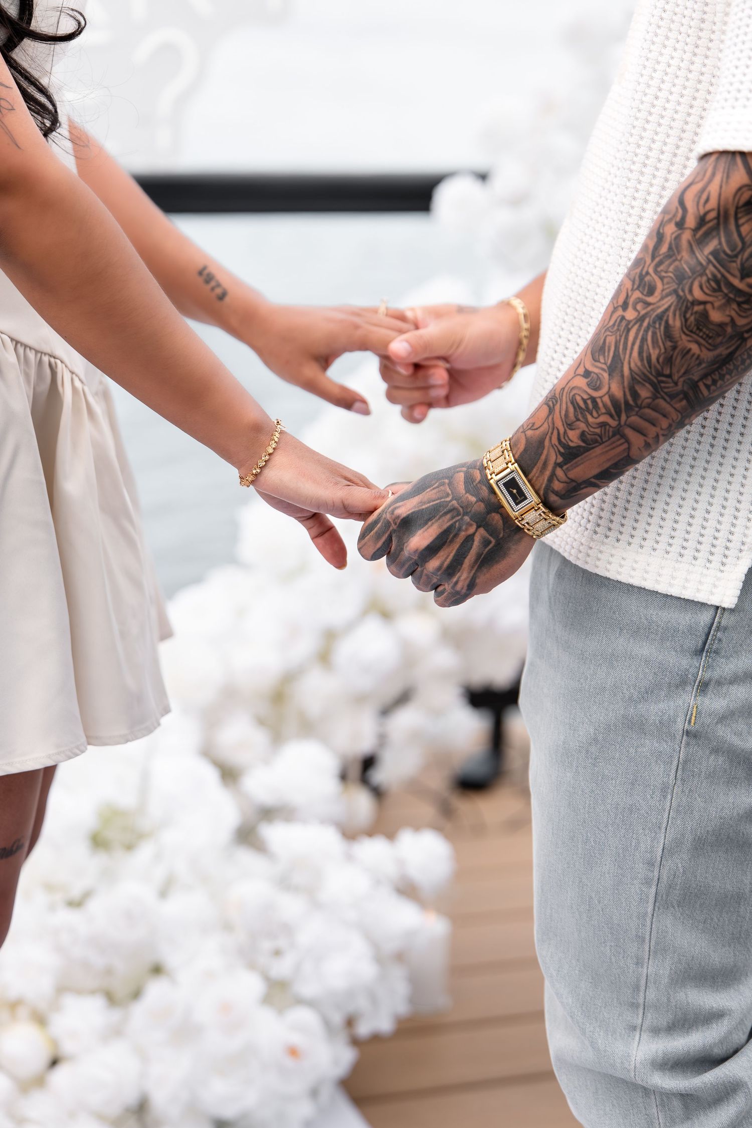 Couple holding hands at a romantic proposal, man with tattoos and gold bracelet, white floral backdrop.
