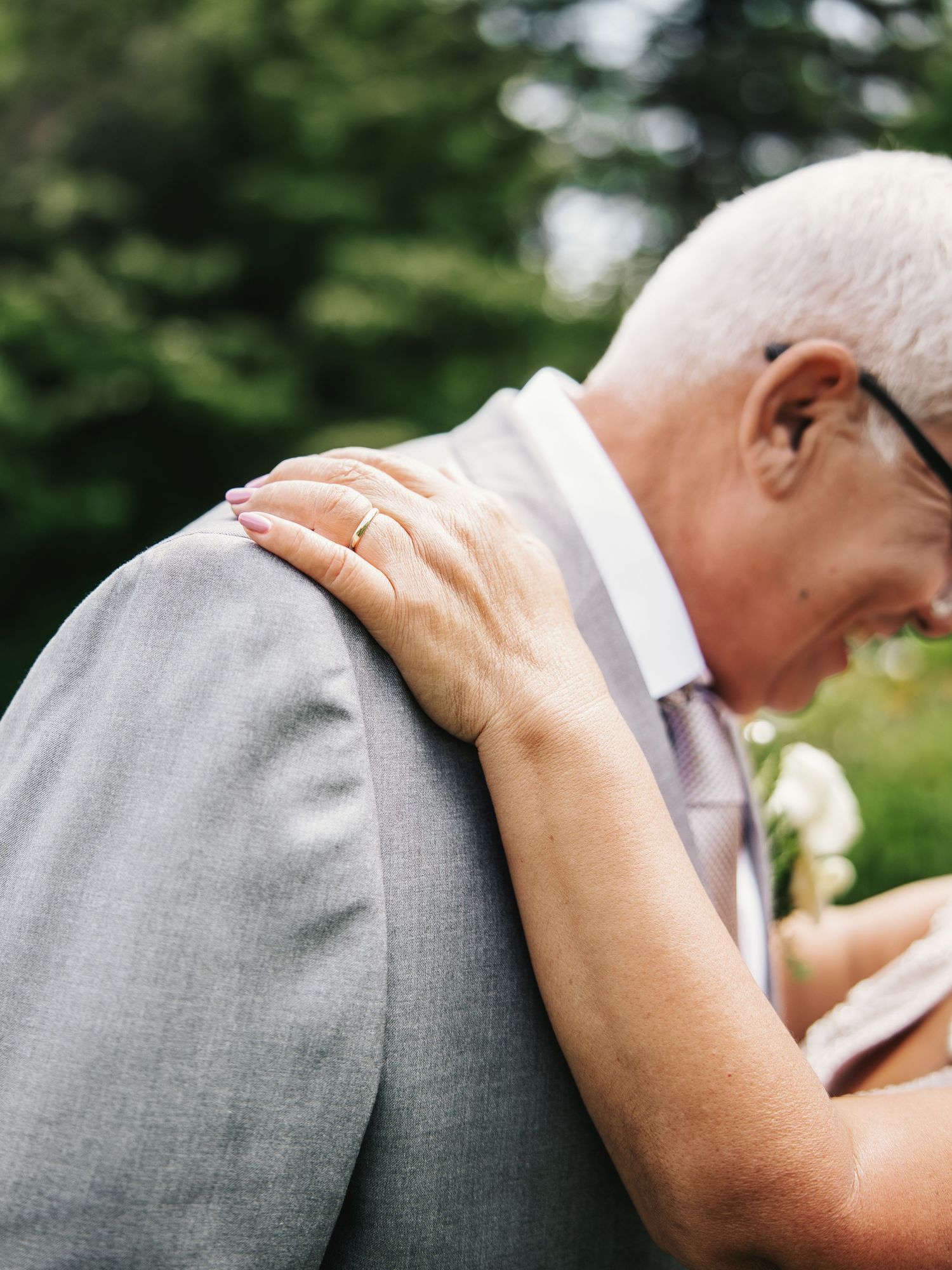 Elderly couple embracing at a wedding, woman's hand with ring resting on man's gray suit shoulder outdoors.