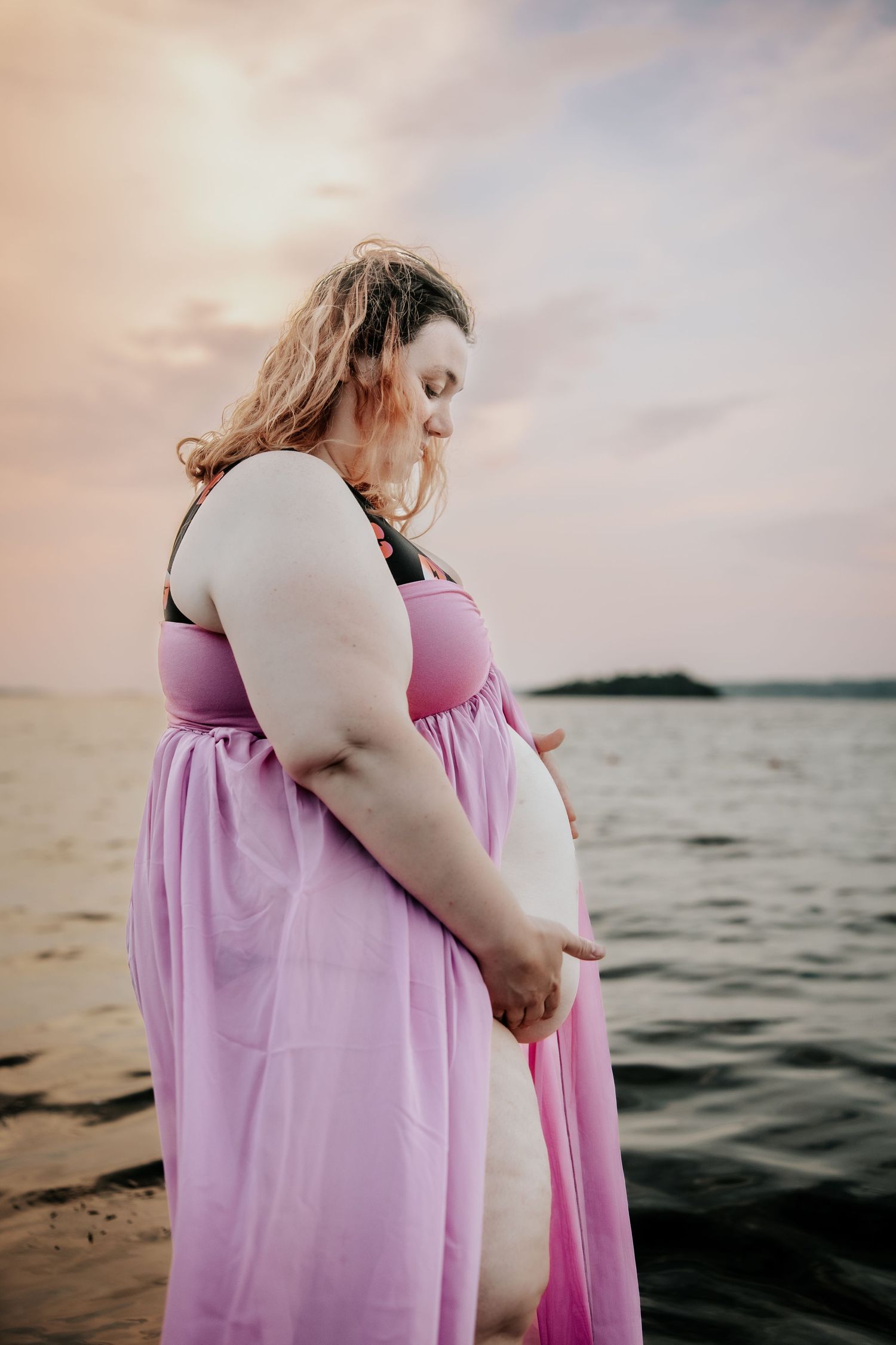 Pregnant woman in pink dress standing by the ocean at sunset, cradling her baby bump.