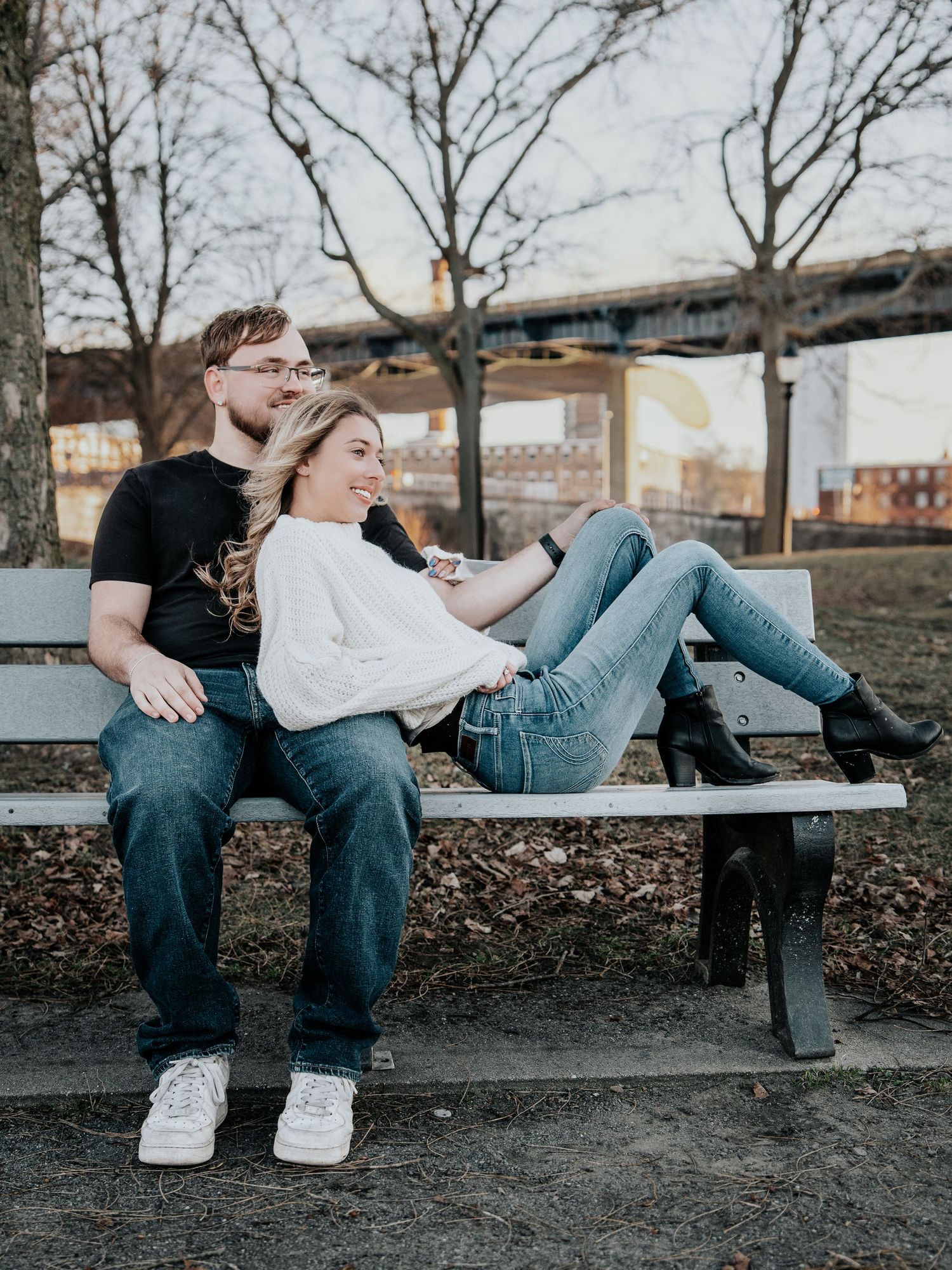 Couple relaxing on a park bench, woman in white sweater resting legs across man in black shirt outdoors.
