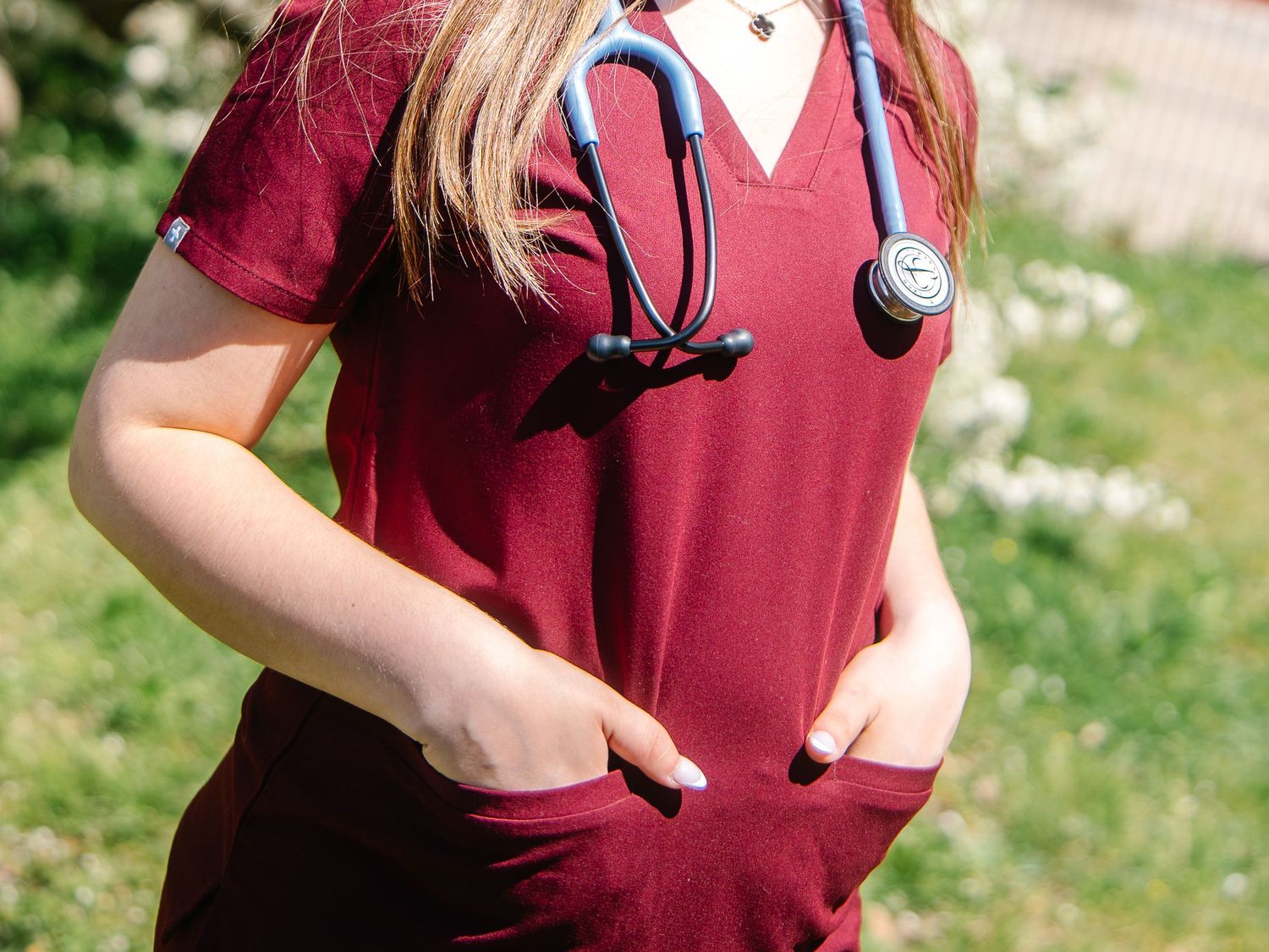 Healthcare student in maroon scrubs with blue stethoscope draped around neck outdoors.
