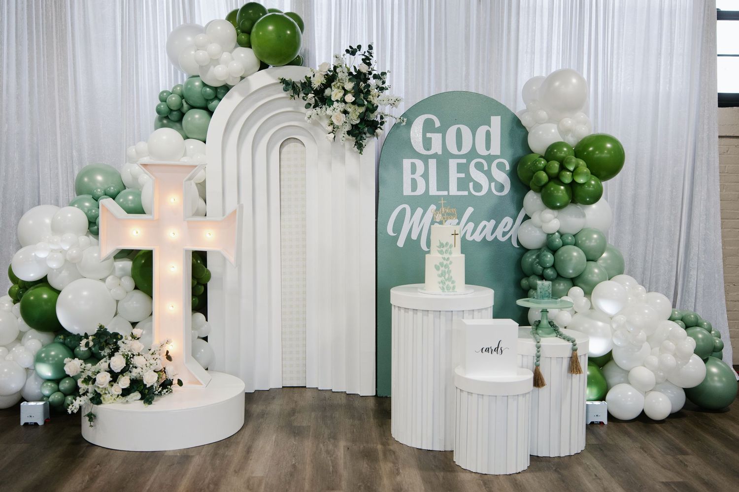 God Bless Michael baptism backdrop with illuminated cross, balloon garland in sage green and white.