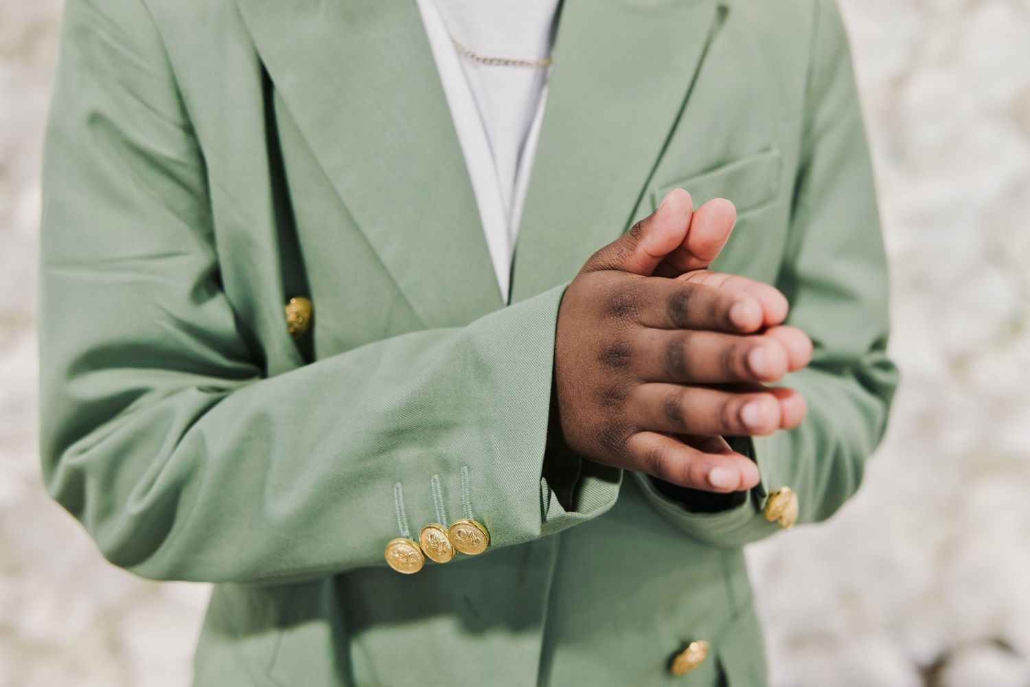 Close-up of a person clasping hands wearing a sage green double-breasted blazer with gold buttons.