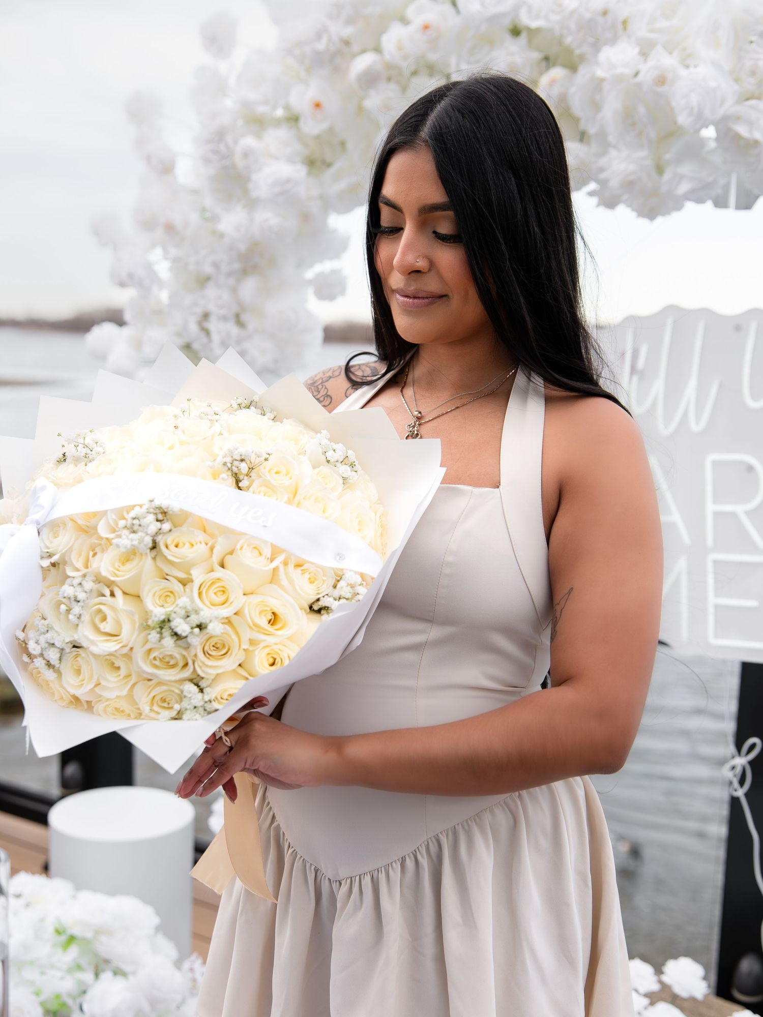 Woman in beige dress smiling while holding a bouquet of white roses at an outdoor romantic event.