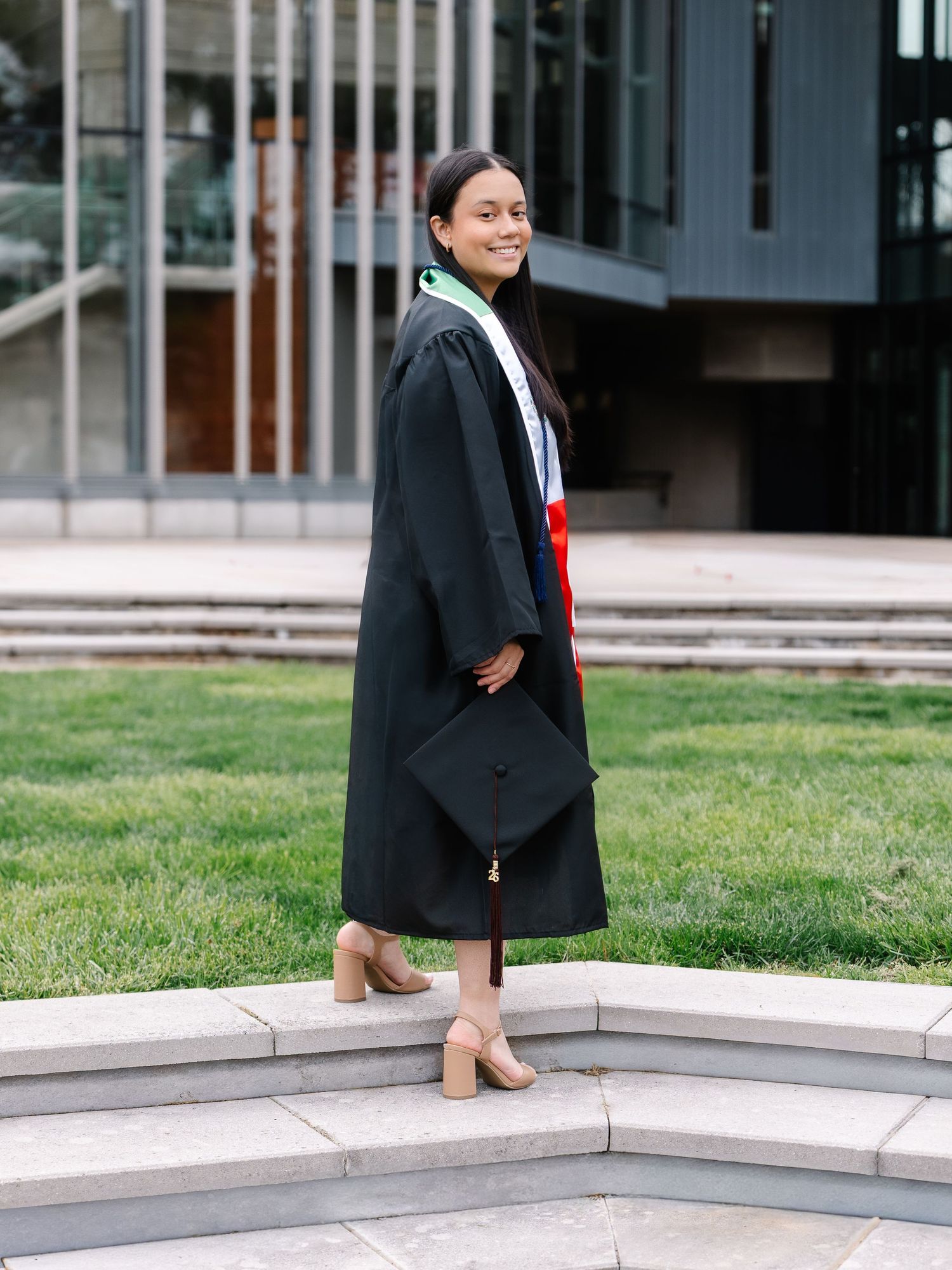 Graduate in black cap and gown holds mortarboard while walking on stone steps outdoors.