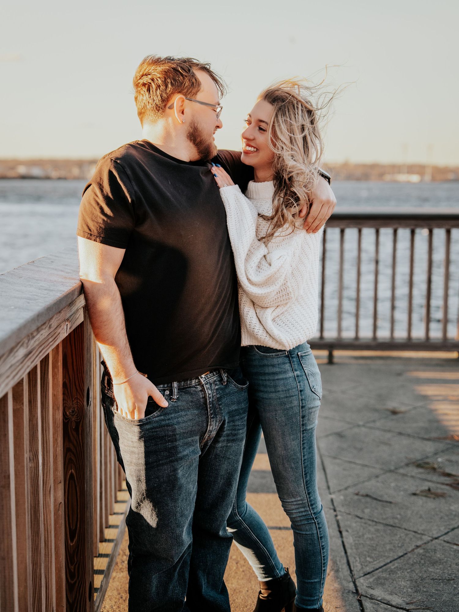 Happy couple embracing by waterfront railing at sunset, woman in white sweater hugging man in black tee.