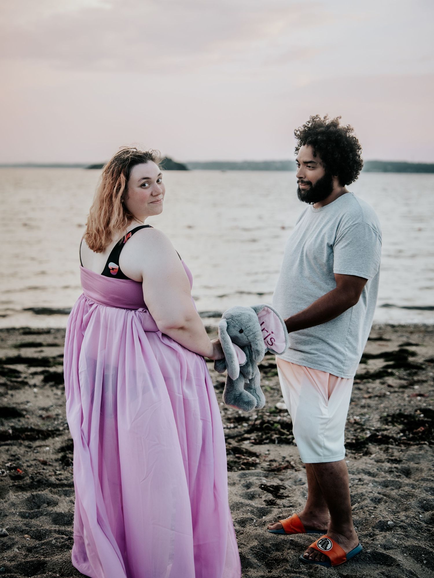 Pregnant couple poses on sandy beach at sunset, woman in flowing pink gown, man holding elephant toy.