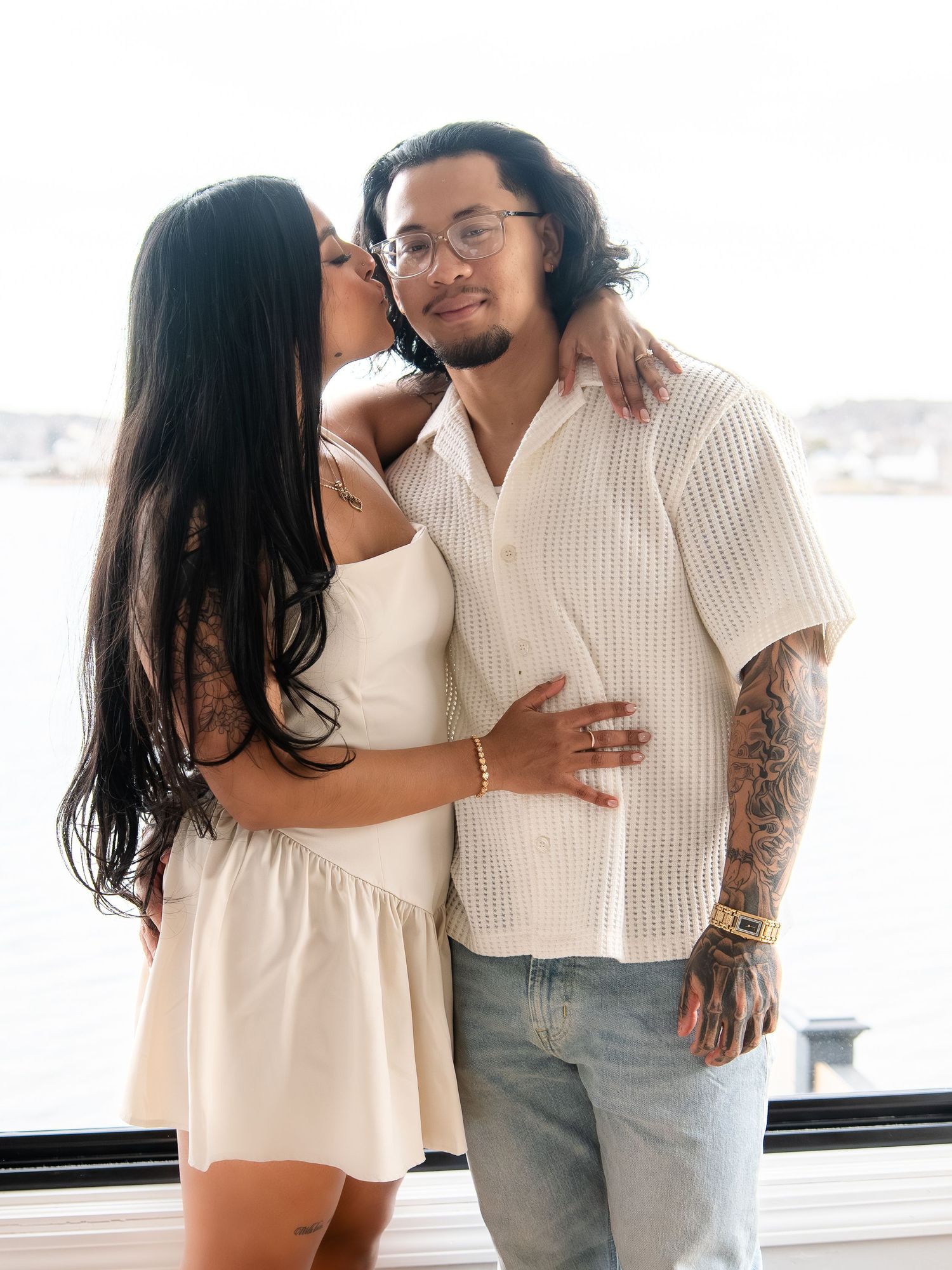 Woman kisses tattooed mans cheek as couple embraces near waterfront window.