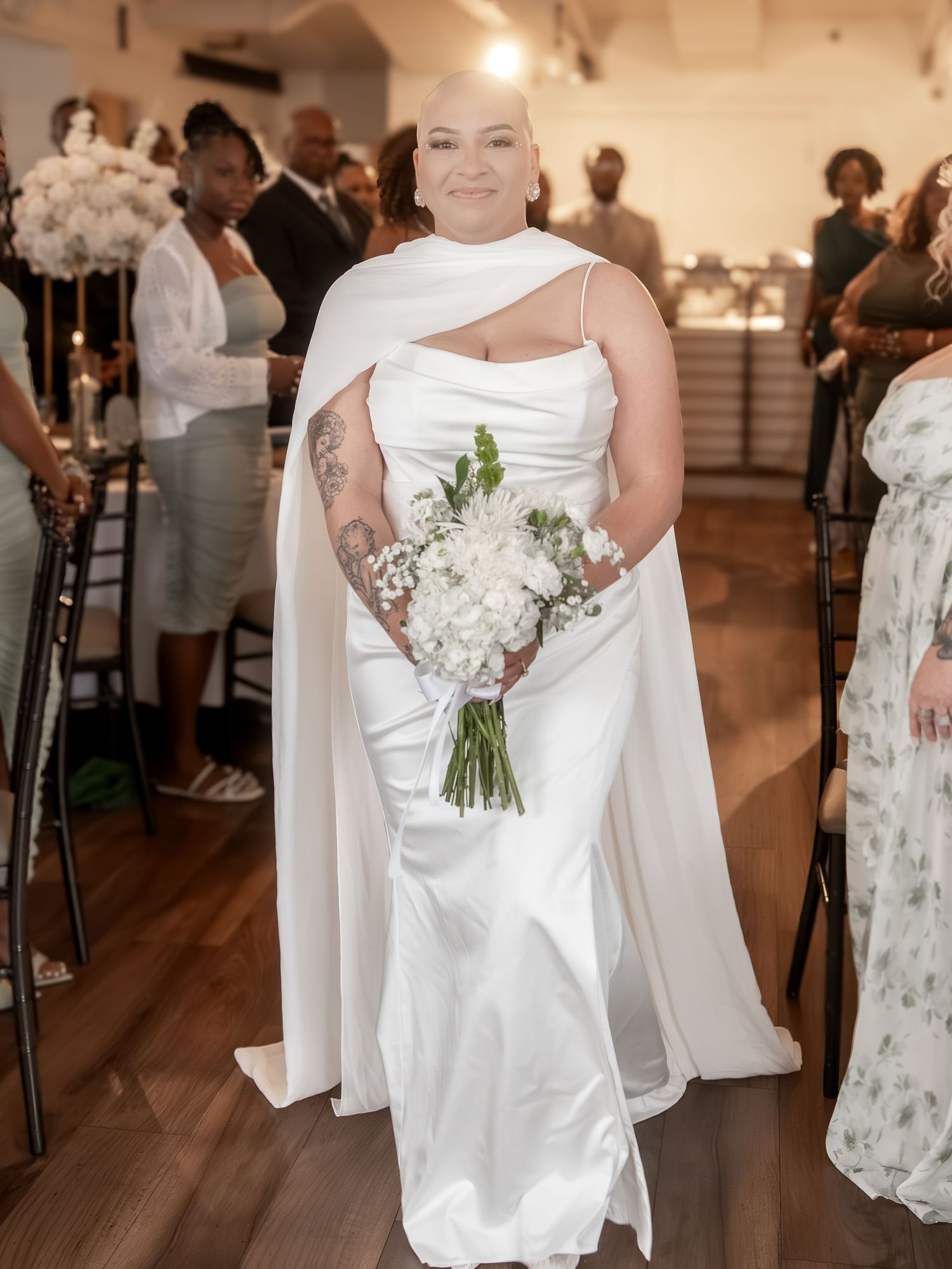Bride in white satin gown with cape walks down aisle holding white floral bouquet at elegant indoor wedding ceremony.