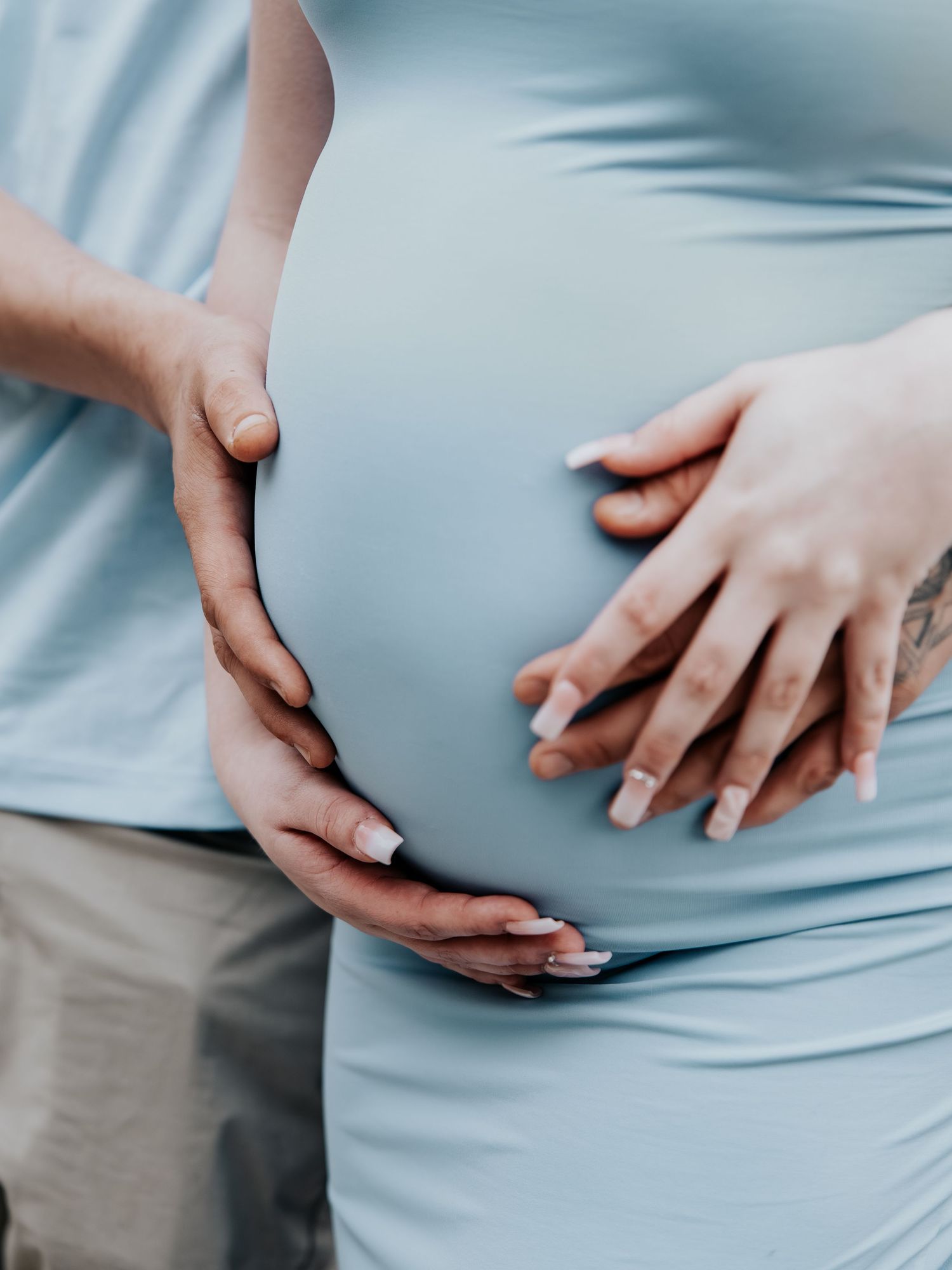 Pregnant woman in light blue shirt with two pairs of hands gently cradling her baby bump.