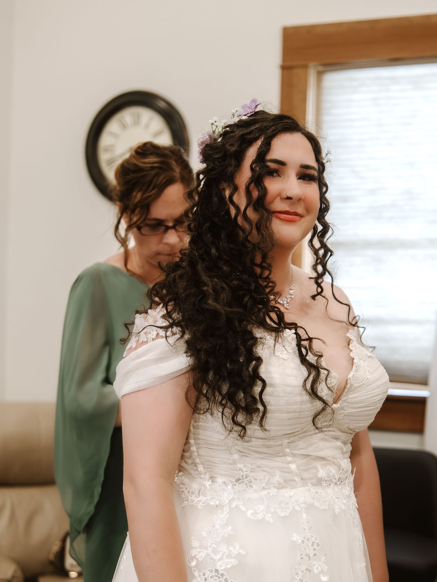 Bride in white lace gown with curly hair gets help from bridesmaid in green dress before wedding ceremony.