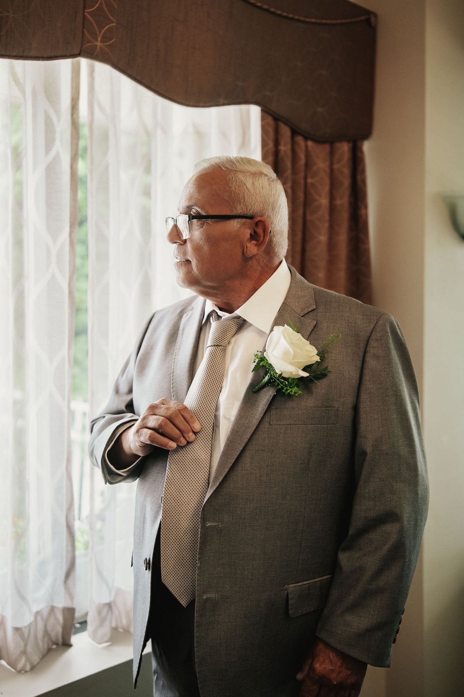Elderly man in gray suit with white rose boutonniere gazes out window, dressed for a formal wedding occasion.